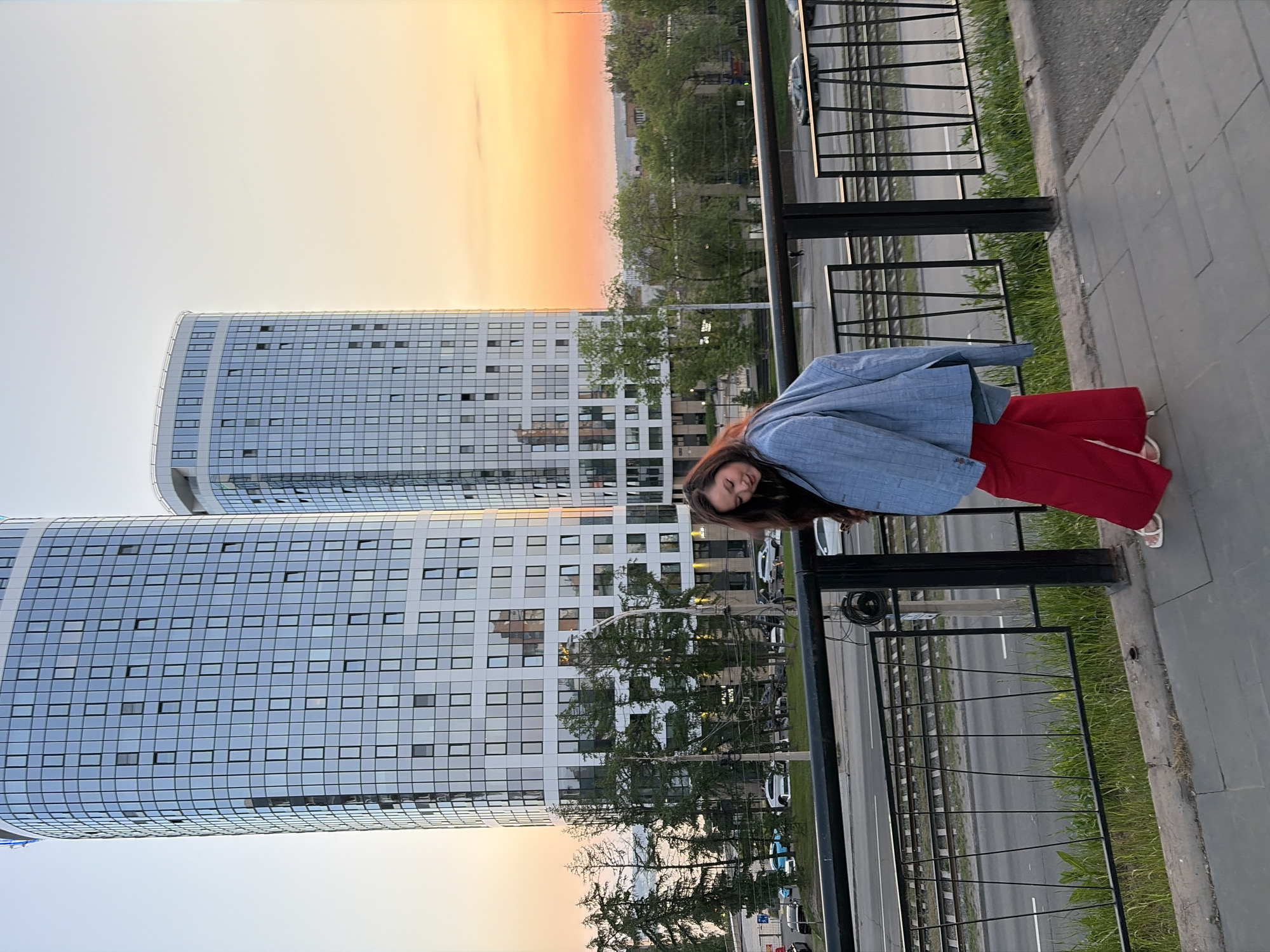 A young woman is posing for a photograph on a sidewalk next to a black metal fence. She is wearing a light blue blazer and red trousers. Behind her are two modern high-rise buildings with glass facades, reflecting the sky. The sky shows signs of dusk, with a mix of orange and yellow hues. The buildings and the fence are aligned along a street with cars visible in the background. The scene appears to be in an urban area, with trees lining the street. The exact location is Izhevsk, Russia.