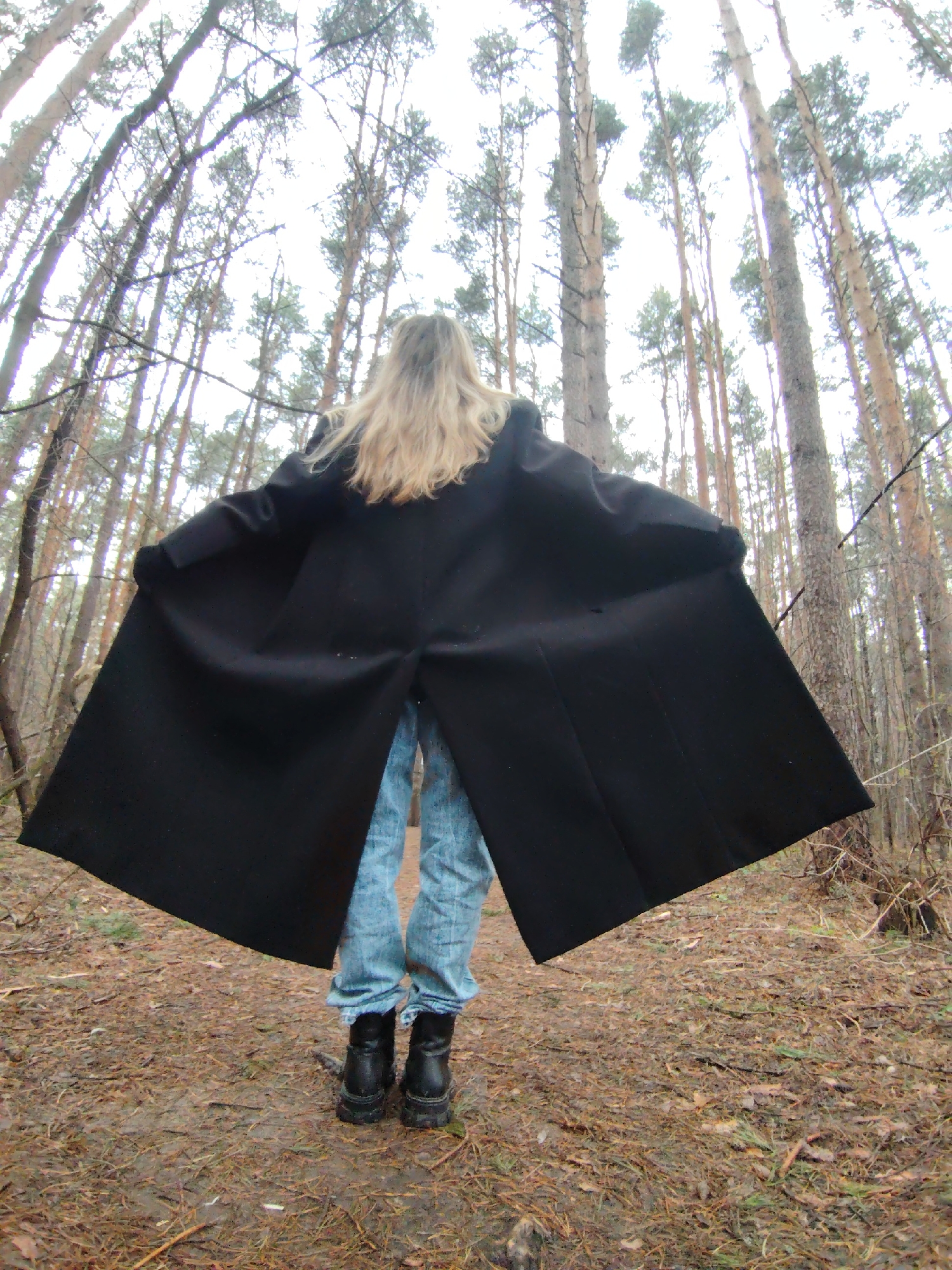A person with long blonde hair, wearing blue jeans and black boots, stands on a path in a forest. They are facing away from the viewer, with their arms outstretched, holding open a black coat. The forest is dense with tall pine trees. The ground is covered in fallen leaves and pine needles. This image was taken in Chaikovsky, Russia.