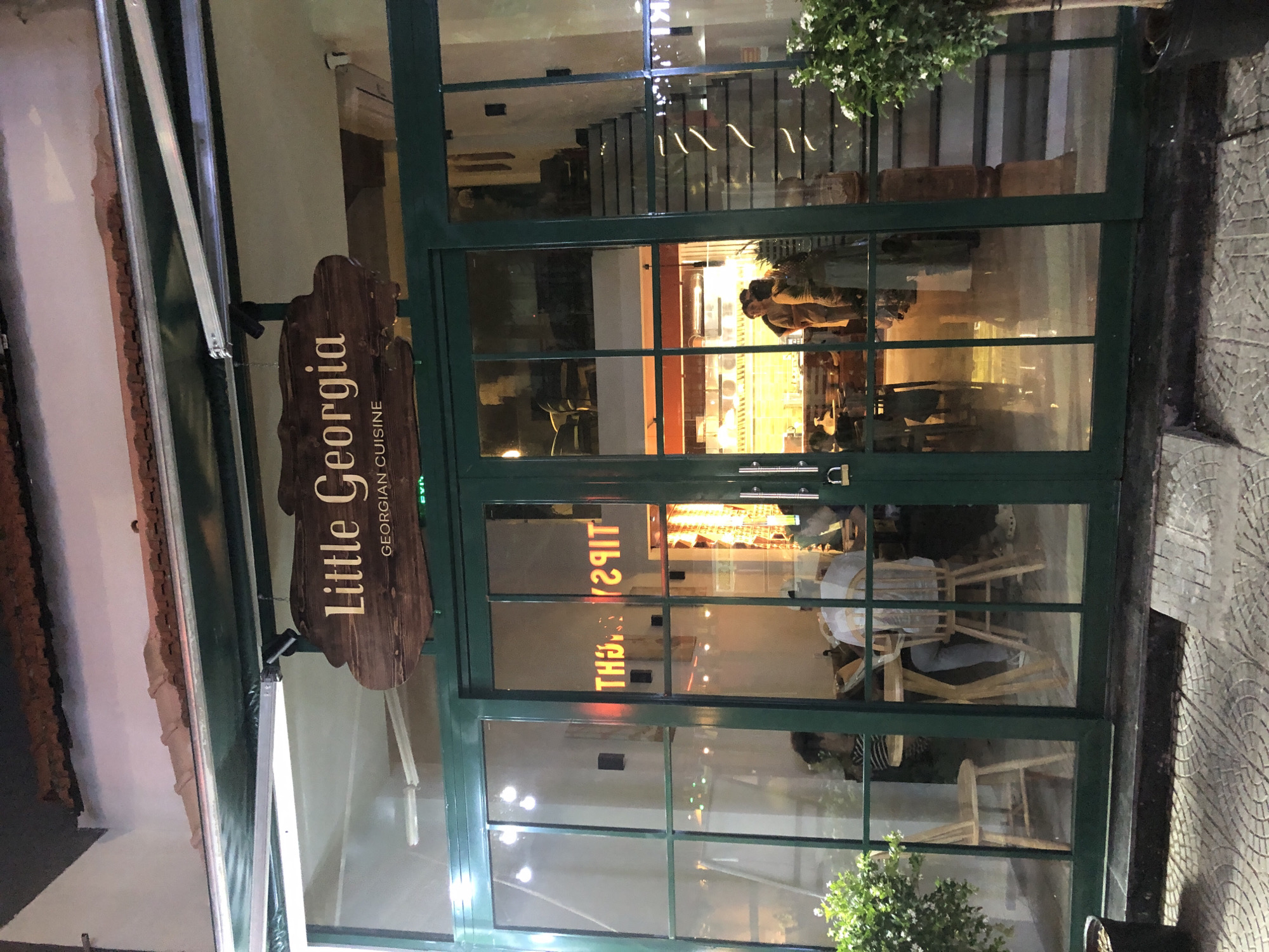 The image shows the exterior of a restaurant named "Little Georgia Georgian Cuisine" located in Đà Nẵng, Vietnam. The restaurant has a green-framed glass facade with a wooden sign above the entrance. Inside, people are visible sitting at tables and walking. The interior is lit by warm lighting, and there is a staircase visible on the right side of the image. The pavement in front of the restaurant is made of patterned tiles.