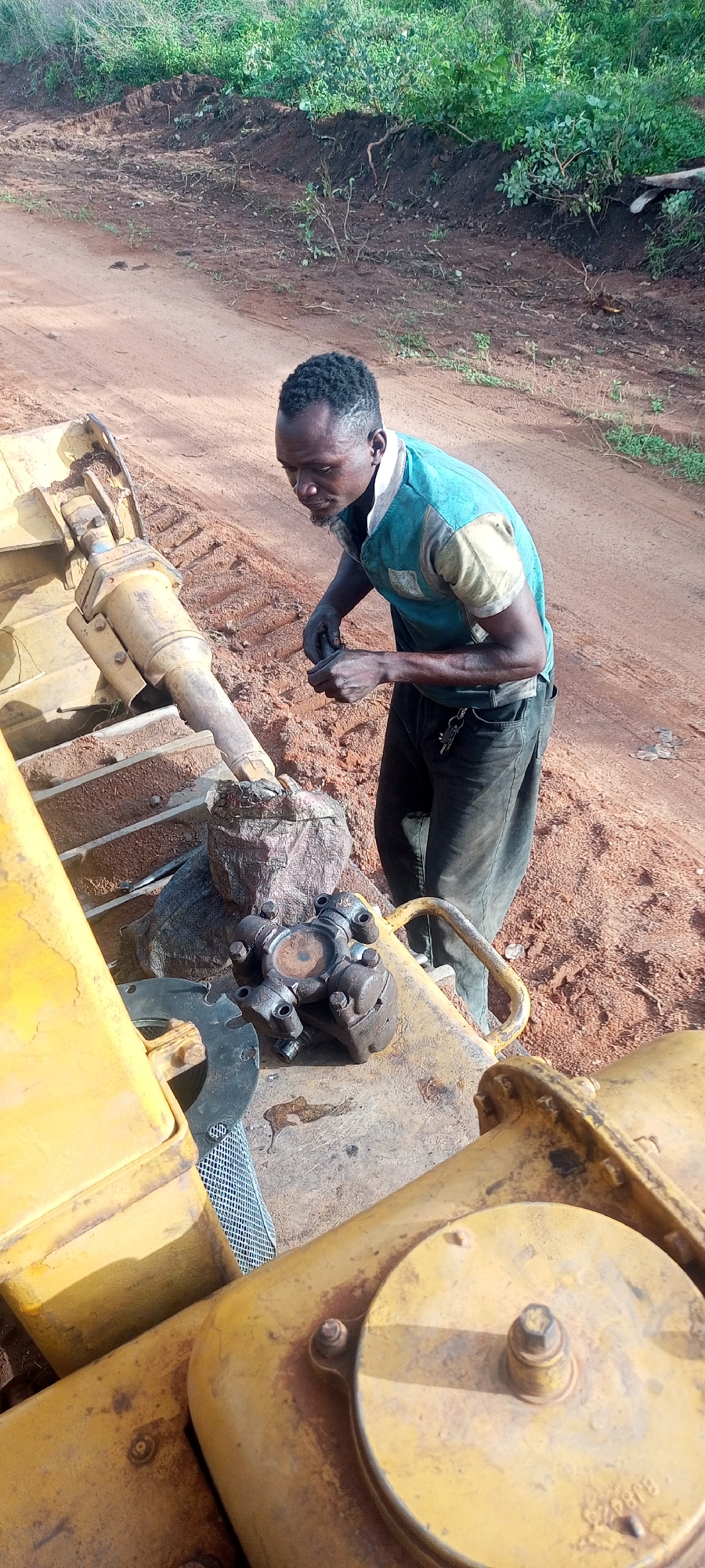 This image captures a focused man performing maintenance or repair work on heavy machinery in an outdoor setting in Otunade, Nigeria.
The central subject is an adult man with short, dark, textured hair and a beard. He is wearing a short-sleeved t-shirt, predominantly teal or light blue with light yellow or cream sections on the shoulders and sleeves, and dark trousers. A set of keys hangs from a loop on his pants. He is leaning over, intently examining or working with his hands on a component of the machinery.
The machinery itself is large and yellow, likely a bulldozer or excavator, evidenced by the visible tracks in the background and a prominent hydraulic cylinder extending from its frame. Resting on a flat surface of the machinery are mechanical parts, including a universal joint (a cross-shaped coupling) and a round metal plate, indicating ongoing mechanical work. On a circular yellow cap towards the bottom right of the frame, the text "BJ926" is faintly visible.
The setting is an unpaved area with reddish-brown earth, which appears to be a typical rural or construction site. In the background, a natural bank of green foliage and disturbed soil completes the scene. The lighting suggests it is daytime, either morning or afternoon, with clear weather conditions. The man's expression is one of concentration and diligence as he works on the equipment.