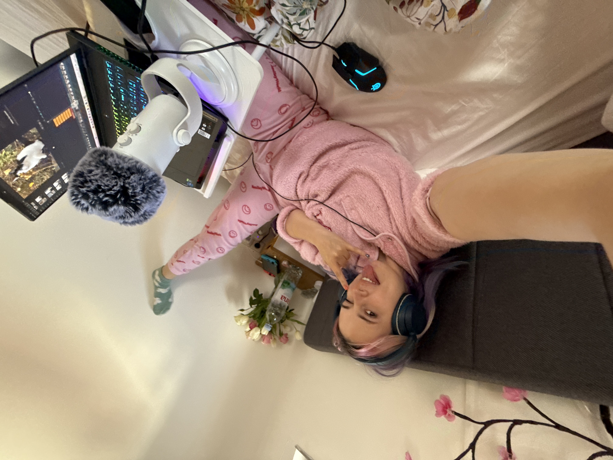 An indoor scene within a residential setting in the City of Zagreb, Croatia, is depicted; the specific location within Zagreb is not identifiable. A young adult female with multicolored hair (pink, purple, blue) is lying on her back on a bed with white sheets and a patterned duvet, propped against a dark grey upholstered headboard. She wears a pink fuzzy long-sleeved top and matching pink pants with red smiley face patterns, along with teal over-ear headphones. Her left arm is extended, holding a device to capture the image. Her right eye is winking, her tongue is extended, and her right hand displays a "rock on" gesture. Her left leg is raised, with her foot, adorned with a green patterned sock, resting against a light-colored wall.
To her right, a wooden bedside table holds a bouquet of white and pink tulips in a glass vase, a bottle of "Jana" brand water, and a blue Nintendo Switch console. Above the bed, an artificial cherry blossom branch with pink flowers is affixed to the wall. Adjacent to the subject on the bed are a black wired gaming mouse with blue LED accents and a white USB microphone featuring a grey furry wind muff and a purple illuminated base. An open black laptop with a blue-green backlit keyboard displays a screen showing what appears to be video editing software, featuring a white bird against a green background. Black cables are visible, connecting the electronic devices. The room has light-colored walls and is well-lit.