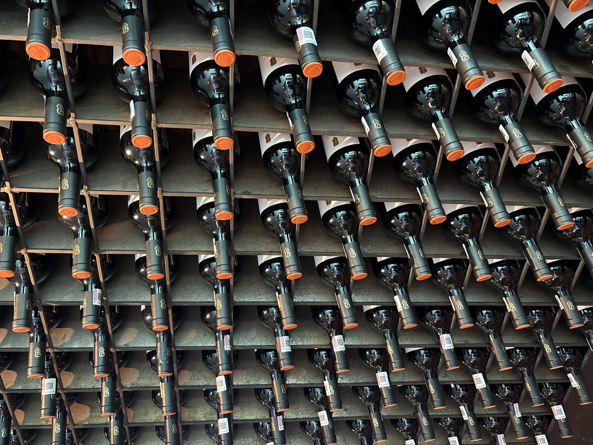 The image shows rows of wine bottles stored upside down in a metal rack. The bottles are dark with orange seals around the bottom. Some bottles have white labels with QR codes attached. The location appears to be a wine cellar or storage area. The image does not depict the city of Huixquilucan de Degollado, Mexico, so I am unable to include location-specific details about that city.
