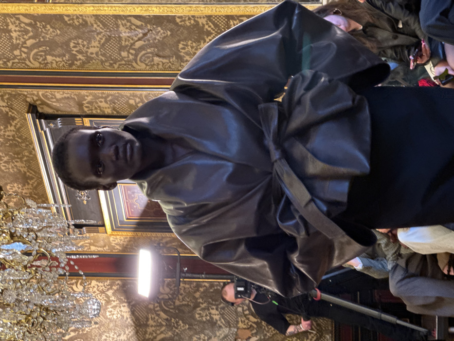 The image depicts a fashion event within an opulent interior in Paris, France, likely a historic salon or private mansion. Centered in the foreground is a dark-skinned individual, appearing to be a model, wearing a voluminous dark, possibly leather, jacket with a wide, structured collar and a prominent tie at the waist, over a black skirt. The model faces slightly right with a neutral expression.
The background features rich gold and brown damask-patterned wallpaper, contrasted with dark wood paneling and gilded accents. A multi-tiered crystal chandelier hangs from the ceiling in the upper left quadrant. To the left, a male videographer or camera operator, dressed in a black t-shirt, stands behind a camera mounted on a tripod. A bright, rectangular LED light panel on a stand is positioned near him, illuminating the scene. Partial views of seated audience members are visible in the lower left and right; one individual in the lower right holds a smartphone.