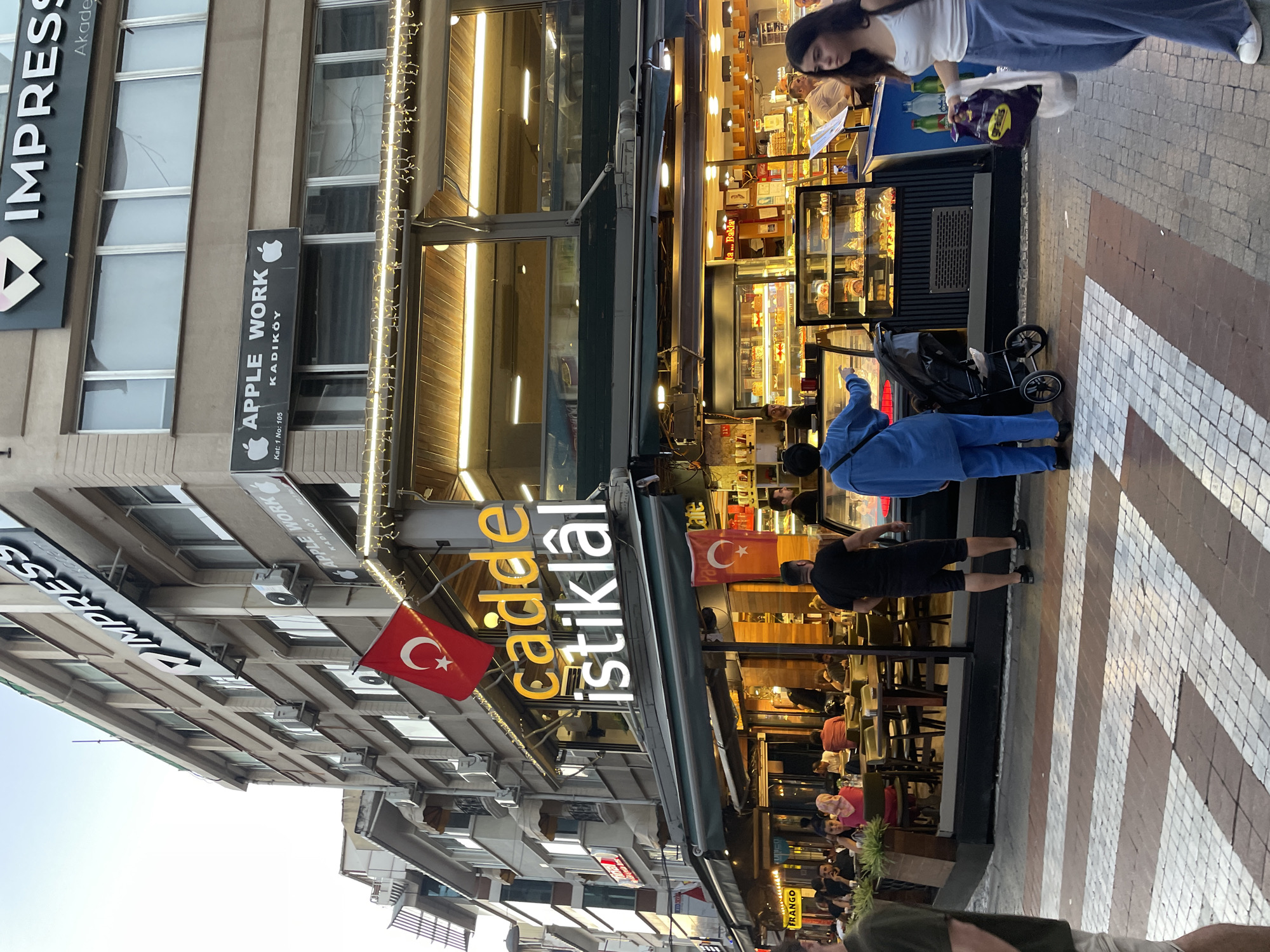 The image depicts a street scene in Kadıköy, Turkey, likely on or near İstiklal Caddesi, given the prominent signage. The foreground features a pedestrian crossing with a white and brown patterned surface. In the middle ground, a restaurant or cafe with large display windows showcases pastries and desserts. Several people are visible, including a man in a blue outfit pushing a stroller, and a woman in a white top and blue pants walking with shopping bags. To the left, patrons are seated at tables outside the establishment. The background reveals a multi-story building with signage for "Impress" and "Apple Work Kadıköy." A Turkish flag is prominently displayed on the building. The overall impression is of a busy urban commercial area during daylight hours.