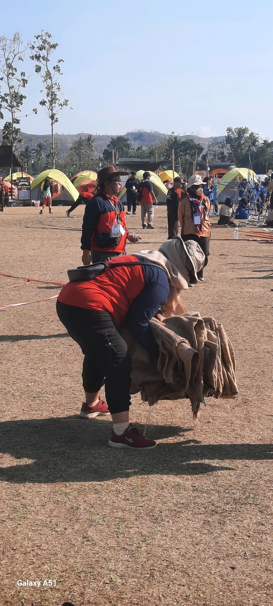 In Selopuro, Indonesia, a group of individuals are participating in an outdoor activity, possibly a scout camp, indicated by the tents and the scout-like attire of some participants. In the foreground, a person wearing a red and blue jacket, black pants, and red sneakers is bent over, holding a bundle of brown fabric. Another person in a red and blue jacket and brown hat stands to the left, facing right, with a lanyard around their neck and a fanny pack. In the background, several colorful tents are set up, and other people are milling about. The environment is an open field with dry grass and scattered vegetation, with hills visible in the distance under a clear blue sky. The lighting suggests it is daytime.