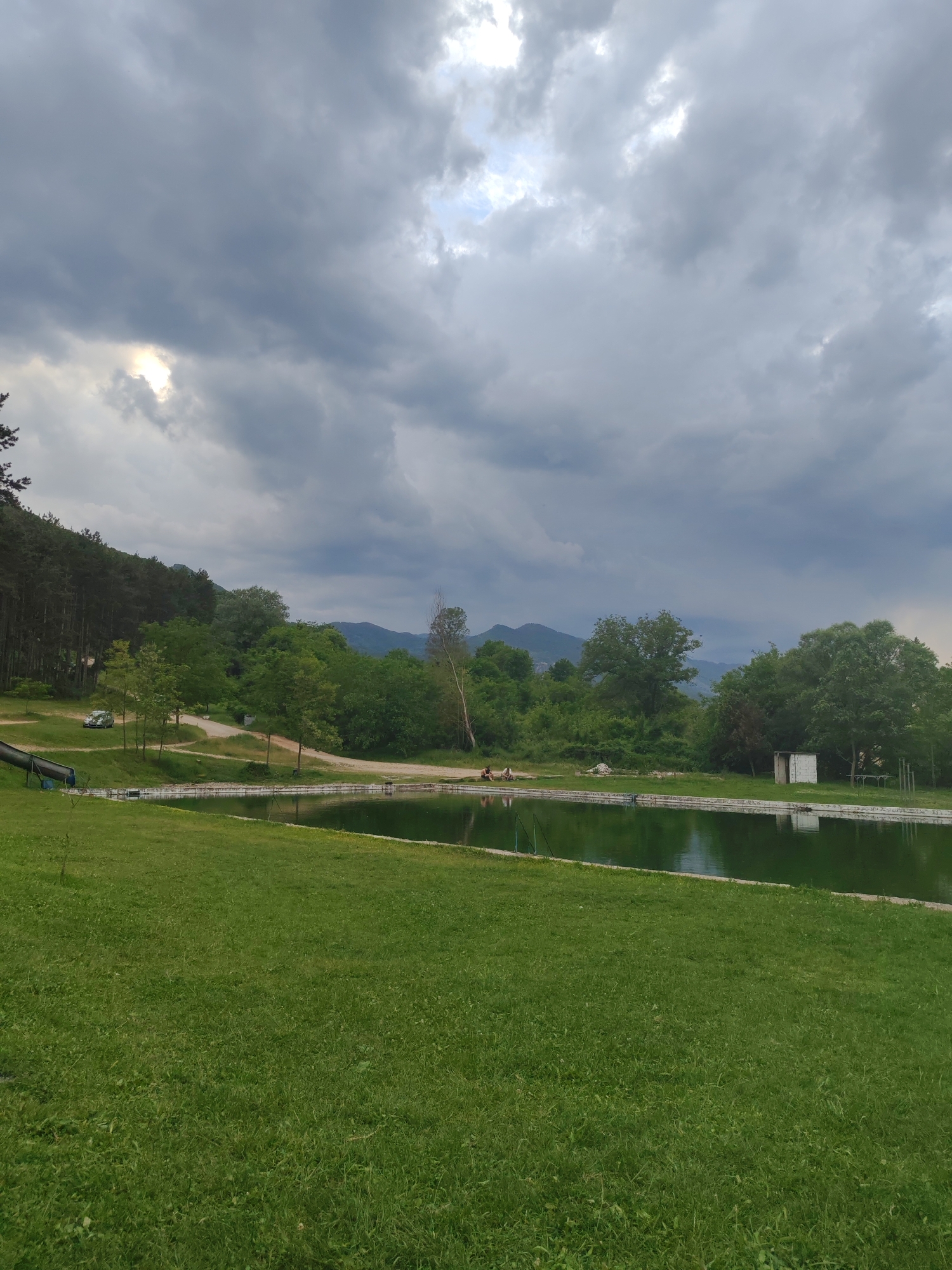 An outdoor scene captures a wide view of a natural or recreational area under a heavily overcast sky. In the foreground, a large expanse of vibrant green grass stretches towards a body of water, likely a pond or small lake, which occupies the middle ground. The water has a greenish hue and reflects the cloudy sky and surrounding vegetation. On the far bank of the water, a dirt path winds through green areas.
Several people are visible in the mid-ground. To the left, a silver car is parked on the dirt path next to a forested hillside. Below the car, a blue water slide descends towards the water, although no one is actively using it. Further along the far bank, towards the right, two separate small groups of people are seated or relaxing by the water's edge. One group is near the center, and another is closer to the right side, next to a small, white, rectangular building or shed. The background features dense green trees and hills, with a range of mountains faintly visible under the distant clouds.
The sky is dominated by thick, dark grey clouds, suggesting an overcast day, possibly before or after rain. Patches of brighter light or sun are breaking through the clouds in the upper left and upper right sections of the sky. The overall impression is one of leisure and nature, with the heavy clouds creating a subdued light. No text is visible in the image.