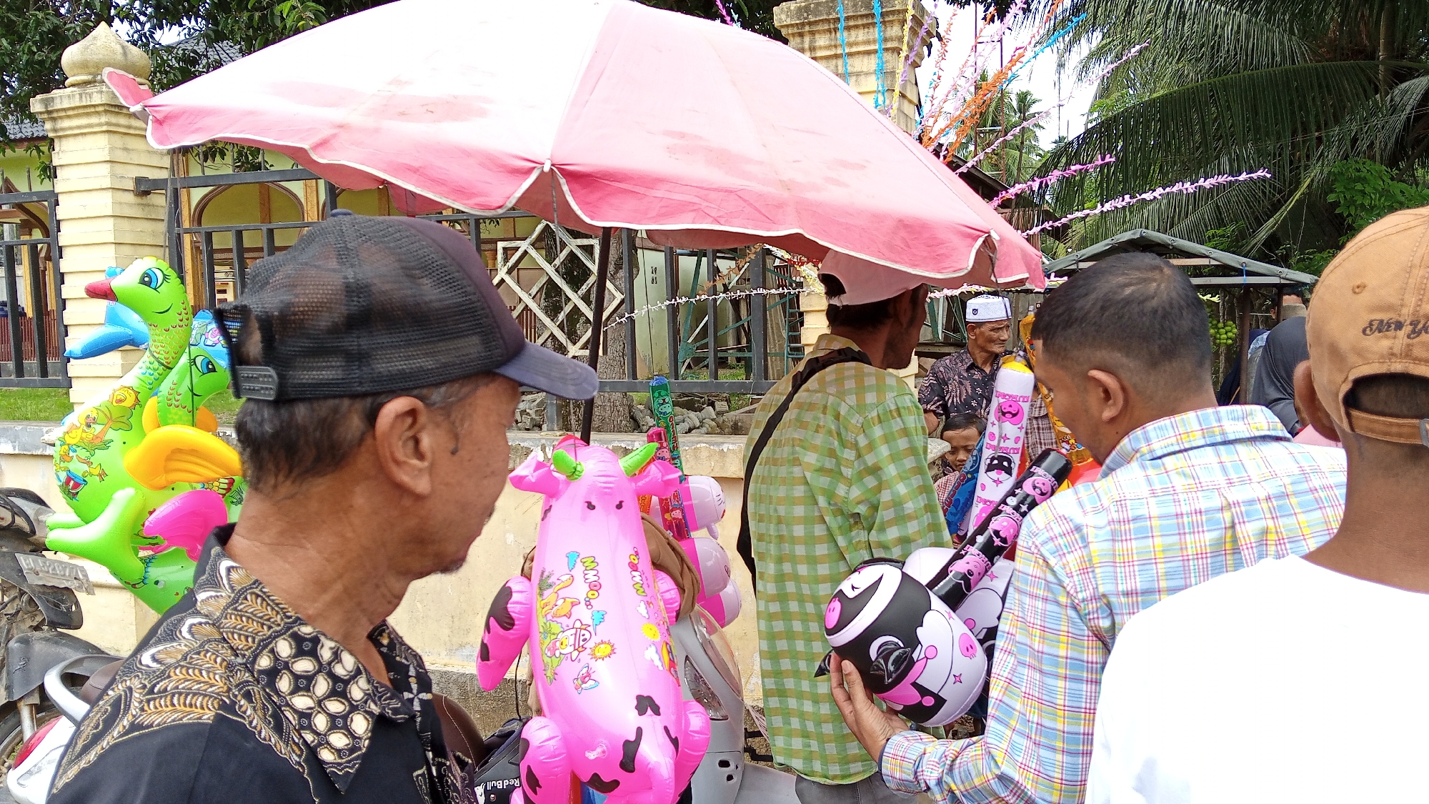 This is a medium shot, eye-level, outdoor photograph taken in Arabungong, Indonesia. The scene depicts a street-level view with several people and inflatable toys displayed under a pink umbrella. In the foreground, a man wearing a black patterned shirt and a dark baseball cap is seen from the side, looking to his left. To his right, a person in a green checkered shirt stands with their back mostly to the camera, holding a pink inflatable cow toy that has "Moo!" written on it. Further to the right, another person, partially obscured, wears a tan baseball cap and a plaid shirt, and is holding a black inflatable toy with a cartoon face.
Behind the main group, an elderly man in a patterned shirt and a white cap is visible, along with a young child. In the background, there is a building with yellow walls and a fence, and lush green trees and palm fronds suggest a tropical environment. Several colorful inflatable toys, including green dinosaurs and pink pigs, are arranged for sale. The overall impression is of a casual outdoor market or street vendor scene in a village.