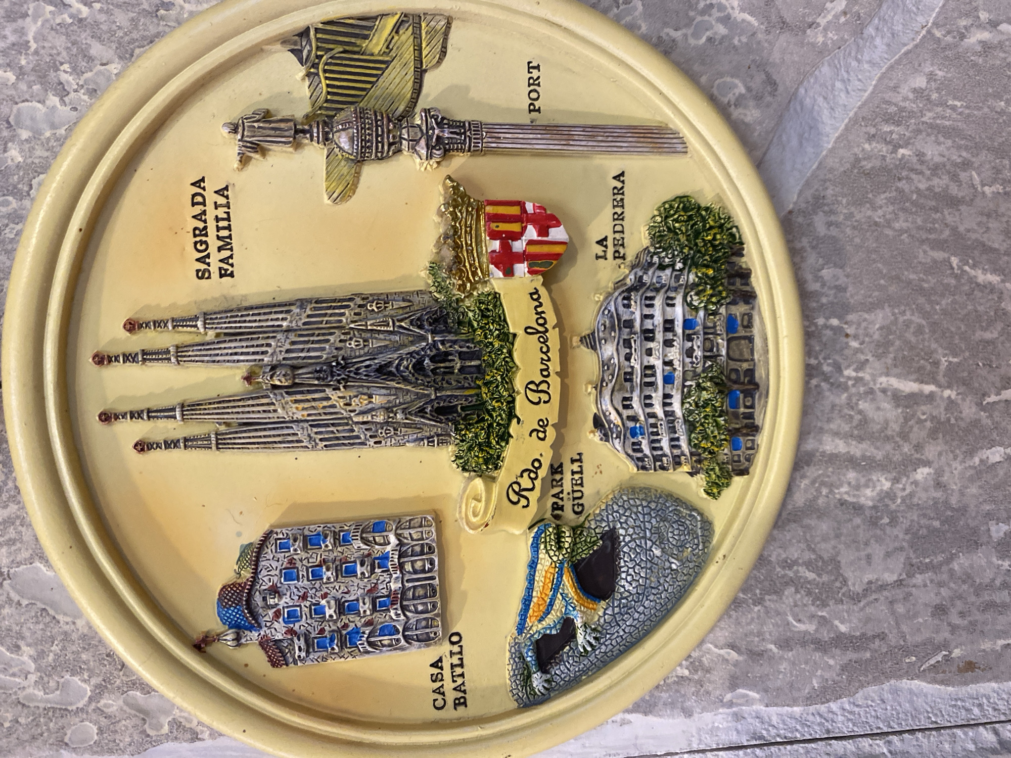 The image is a close-up of a circular, textured souvenir magnet depicting various landmarks from Barcelona, Spain. The magnet is mounted on a grey, stone-like surface. The magnet itself is a pale yellow color with a raised, circular rim.
Prominently featured in the center is a miniature, gothic-style model of the Sagrada Familia basilica, with intricate spires and architectural details. To the left of the Sagrada Familia, a small model of Casa Batlló, a colorful, wavy facade building, is visible. Below Casa Batlló is a mosaic-like sculpture of a lizard, a characteristic element of Park Güell. Text labels around these landmarks identify them as "CASA BATLLO", "PARK GÜELL", and "SAGRADA FAMILIA".
To the right of the Sagrada Familia, a miniature column with a statue on top stands, and further to the right is a section of what appears to be the Barcelona port, labeled "PORT". A shield with a coat of arms, featuring red and yellow stripes and a cross, is positioned between the Sagrada Familia and a building labeled "LA PEDRERA". Another building, "LA PEDRERA", is depicted at the bottom center of the magnet. A ribbon banner across the bottom reads "Rdo. de Barcelona".
The overall impression is that of a tourist souvenir magnet showcasing key attractions of Barcelona. The image does not depict any people or actions. The environment is a plain, textured background that appears to be a wall or surface for display. There is no indication of the location being in Novaya Usman', Russia.