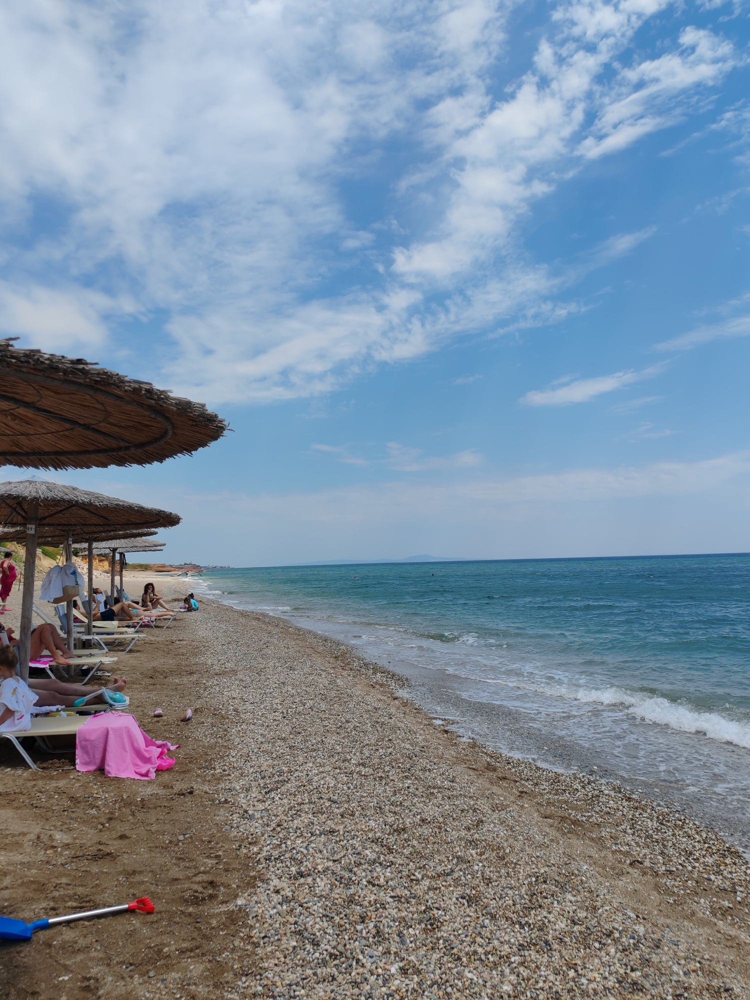 The image depicts a scenic beach with a view of the turquoise sea under a bright blue sky scattered with fluffy white clouds. Several thatched umbrellas provide shade for beachgoers relaxing on lounge chairs. Some people are visible on the beach, either sitting or reclining under the umbrellas. A pink towel is spread out on the sand, and a blue shovel with a red handle lies in the foreground. The beach itself is a mix of sand and pebbles, with gentle waves lapping at the shore. The scene suggests a pleasant day at the beach, likely in the afternoon, given the bright sunlight and clear skies. The location appears to be a tranquil coastal area, possibly a resort or a popular spot for recreation. There is no visible text in the image.