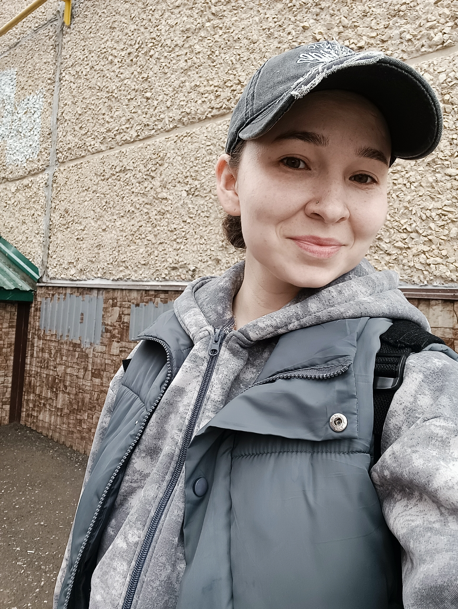 A young woman poses for a selfie outdoors. She wears a gray baseball cap, a gray hooded sweatshirt, and a gray puffy vest. She has a nose piercing on the left side. The background is a light brown concrete building with gray accents. A green awning is partially visible on the lower left side of the image. The setting could be Izhevsk, Russia based on context, but specific location details are not discernible in this image.