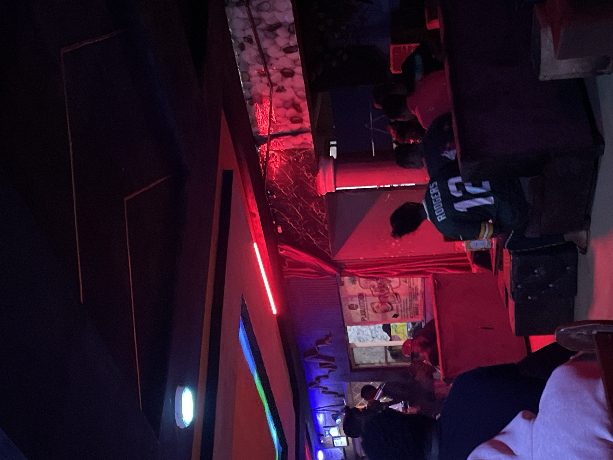 The image depicts an indoor scene, possibly a nightclub or a lounge, in Durumi, Nigeria. The environment is dimly lit, with vibrant red and blue neon lights illuminating the space. Several individuals are present, most of whom are seen from behind or in partial profile. In the foreground, a person wearing a pink hoodie is partially visible, and to their right, another individual is seated, wearing a green and yellow football jersey with the name "RODGERS" and the number "12" on the back. Further into the scene, other patrons are seated and conversing. The decor appears to include textured walls and curtains, with posters or signage visible on one wall. The overall atmosphere suggests a social gathering or entertainment venue.