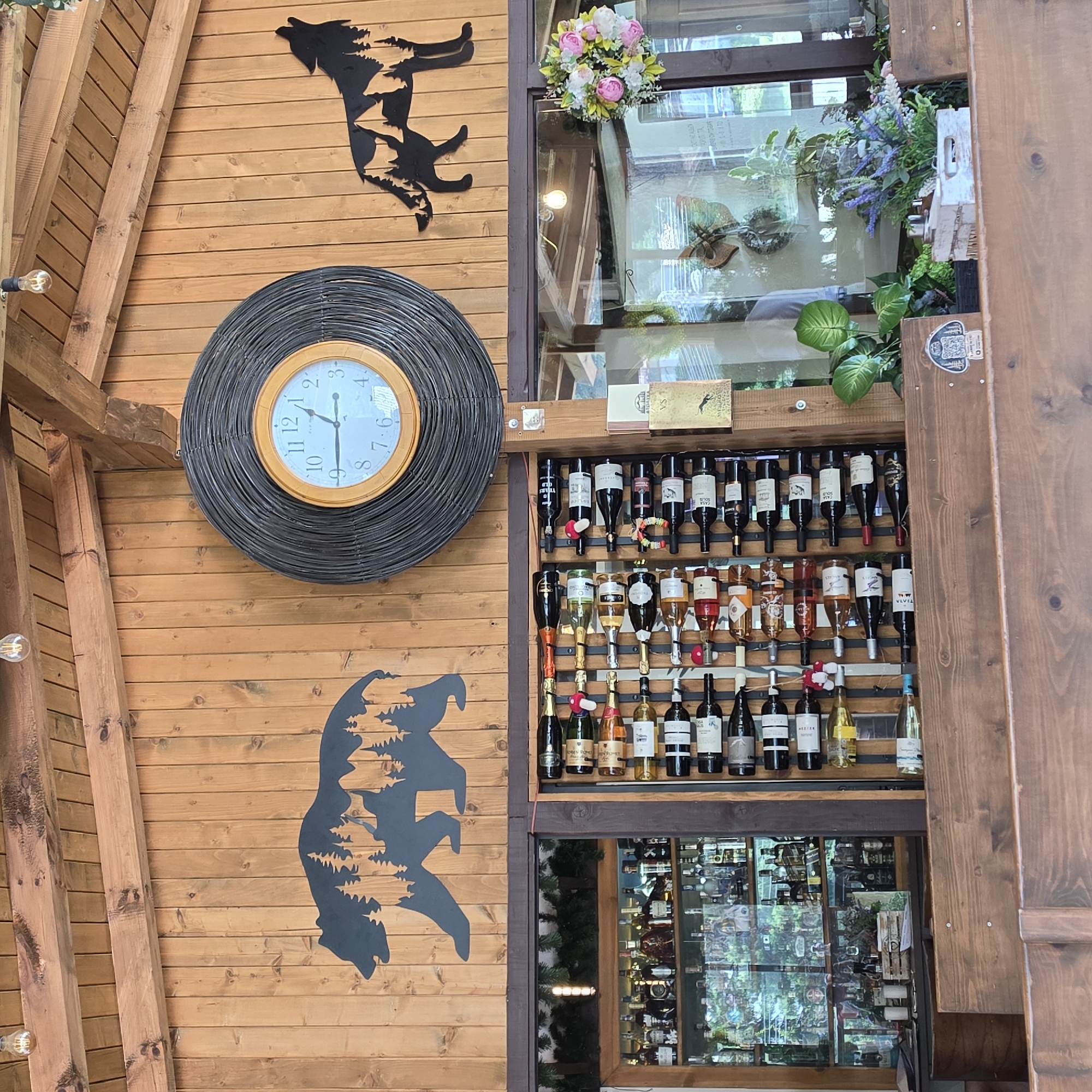 The image depicts an interior scene at a location identified as "Atoluka, Bulgaria." The environment is characterized by exposed wooden beams and vertical wooden paneling on the walls and ceiling. On the wall, there are two silhouette art pieces: one of a bear with trees inside its silhouette on the left, and another of a wolf howling with trees inside its silhouette on the right. A large, circular wicker-framed clock with a white face and black Roman numerals is centrally located between the two silhouettes.
Below the wall art, a section of the establishment features a large glass-fronted cabinet filled with various bottles of liquor and spirits. Adjacent to this cabinet is a wall-mounted wine rack holding numerous bottles of wine. Some of the wine bottles have decorative elements, such as small mushroom-shaped ornaments.
In the foreground, a wooden counter or bar area is visible. The overall impression is that of a rustic-themed establishment, possibly a bar, restaurant, or winery, with decorative elements that evoke a natural or wilderness theme. No people are visible in the image.