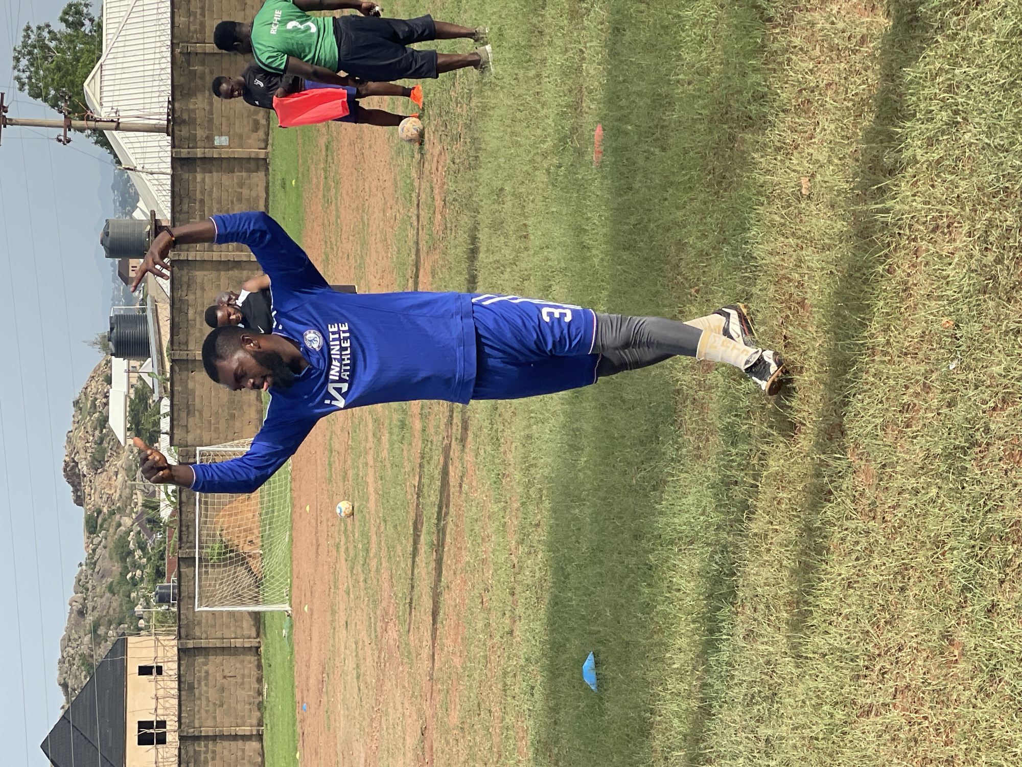 On an outdoor field with sparse, mixed-green and brown grass, a man wearing a blue soccer jersey and shorts, with gray leggings, is the central figure. The jersey has a logo that reads "INFINITE ATHLETE" and the number "3" on the shorts. He stands with his arms outstretched, pointing with both index fingers, his gaze directed downwards. Behind him, there is a small soccer goal with a white net. In the background, two additional individuals are visible, one wearing a green jersey with black shorts and the number "3" on their back, the other's attire is partially obscured. A soccer ball is near them. In the distance is a partly constructed building with black roofs. There are also water tanks, power lines, and a large rock formation with sparse vegetation in the far background. The location is Jos, Nigeria.