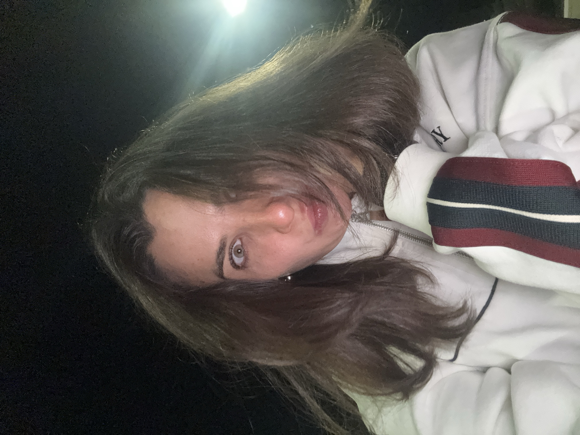 The image presents a rotated close-up portrait of a person, likely female, in a dimly lit setting, possibly in Tivat, Montenegro. The subject has long, dark brown hair, with some strands appearing lighter due to the lighting. One eye, a distinctive brown-hazel color, is clearly visible and looks directly at the viewer with a neutral and direct expression. Parts of the person's nose and lips are also visible.
The individual is wearing a white zipped jacket or sweatshirt. A prominent detail of the attire is the ribbed cuff on the right side of the image, featuring horizontal stripes in dark red (burgundy), dark blue (navy), and white. On the white fabric of the jacket, a partial word or logo is visible, with the letter "Y" clearly discernable, suggesting part of a brand name. The zipper of the jacket is also partially visible.
The background is predominantly dark black, indicating nighttime or a very low-light environment. A strong, artificial light source in the upper left corner creates a bright flare, illuminating portions of the subject's hair and casting some sheen on the skin. The overall impression is a casual, direct photo taken in low light.