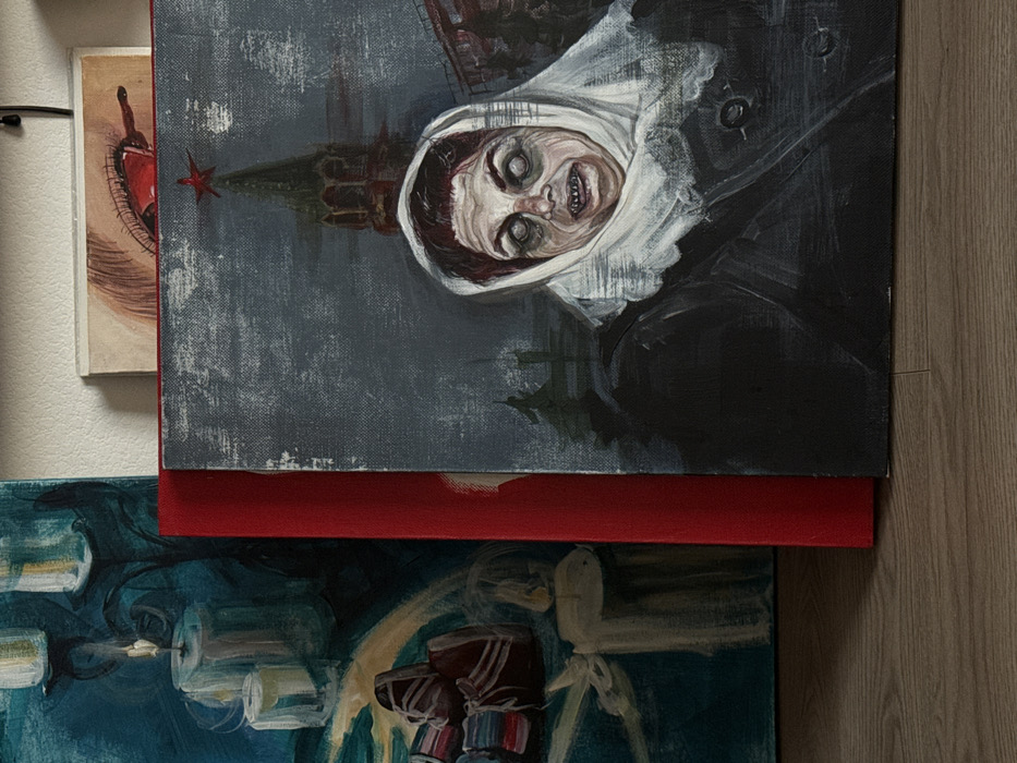 Several paintings are displayed upright on a light brown wooden floor, leaning against a white wall.
The prominent painting in the foreground on the right depicts a grotesque female figure from the chest up. She wears a white headscarf that covers her hair and frames her face, and a dark coat with visible buttons. Her face is pale with a yellowish tint; her eyes are wide, white, and vacant, and her mouth is open, revealing discolored teeth and gums, suggesting an expression of distress or horror. In the background, a distinct red-roofed tower, characteristic of the Moscow Kremlin (e.g., Spasskaya Tower), is visible in the upper center, topped with a red star. This architecture places the depicted scene in Moscow, Russia. The background is a somber grey with dark, indistinct architectural or natural shapes.
To the left, another painting is partially visible, featuring a dark teal background. It portrays multiple lit white candles on a dark, ornate candelabra, with a glow emanating around them. Below the candles, indistinct, stylized shapes are depicted.
Behind the prominent painting and above a red-bound canvas or book spine, a small portion of a third artwork is visible. This segment shows a detailed depiction of an eye with a reddened sclera and red liquid, possibly blood or tears, dripping from the inner corner.
All artworks are resting on a light brown, wood-patterned floor, with a plain white wall serving as the backdrop.