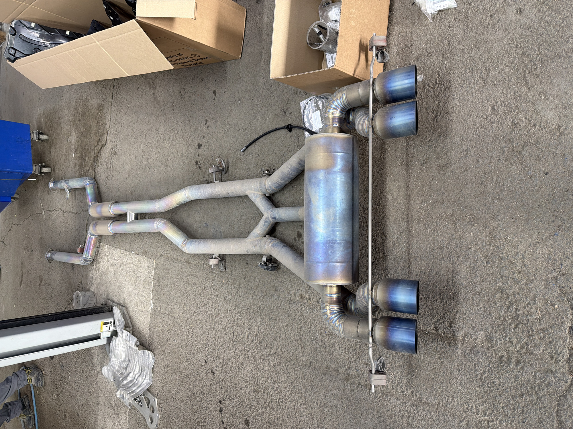 An aftermarket car exhaust system, likely made of titanium due to its distinctive blue, purple, and gold heat-treated coloration, is laid out on a dusty concrete floor in what appears to be an auto repair shop or garage. The exhaust features a central muffler, complex piping, and four prominent, heat-blued exhaust tips (two on each side). Various hangers and a small black wire (possibly for a sensor) are attached to the system.
In the background, two open cardboard boxes are visible. One box has text "PROPERTY OF HAGEL" and "MADE IN SLOVAKIA" printed on its side, suggesting the origin of the parts. Another box contains various smaller items, possibly components or packaging materials, with a small plastic bag holding an item that has a label reading "K-022.01.00" (partially visible).
To the left, a portion of a blue car lift component is visible, along with a person's feet and lower legs wearing hiking boots and grey trousers, suggesting a worker is present. Near the person's feet, a crumpled silver heat shield or bracket lies on the ground. The concrete floor shows cracks and stains, typical of a workshop environment. The lighting suggests an indoor setting. The scene captures the unboxing or preparation of a performance exhaust system, likely for installation on a vehicle.