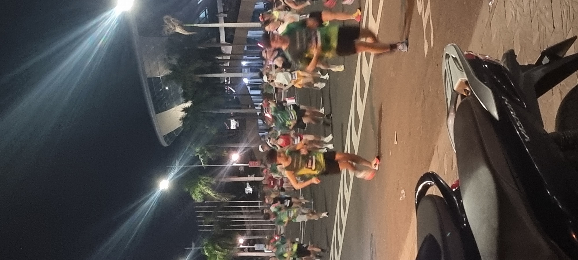 The image depicts a nighttime marathon event taking place on a street in Da Nang, Vietnam. Several runners, predominantly young adults, are visible in mid-stride, wearing athletic attire including t-shirts, shorts, and running shoes. Many are wearing bibs or race numbers, suggesting participation in an organized competition. The street is illuminated by artificial lighting, including streetlights and spotlights, casting distinct beams across the scene. Palm trees line the background, with a large, modern building, possibly a stadium or arena, visible in the upper right quadrant of the image. In the foreground, a parked motorcycle is partially visible, occupying the bottom portion of the frame. The overall atmosphere suggests a lively sporting event occurring in an urban setting.