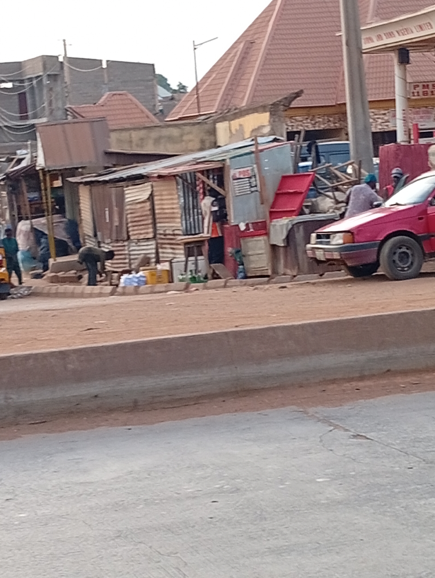 The image depicts a street scene in Nassarawa, Nigeria. There are several small, makeshift shops constructed from corrugated iron and other materials lining the roadside. People are present, some appearing to be engaged in activities near the shops. A red car is parked near one of the structures. In the background, there are buildings with reddish-brown roofs, and a gas station is also visible. The road is visible in the foreground and appears to be made of concrete or asphalt.
