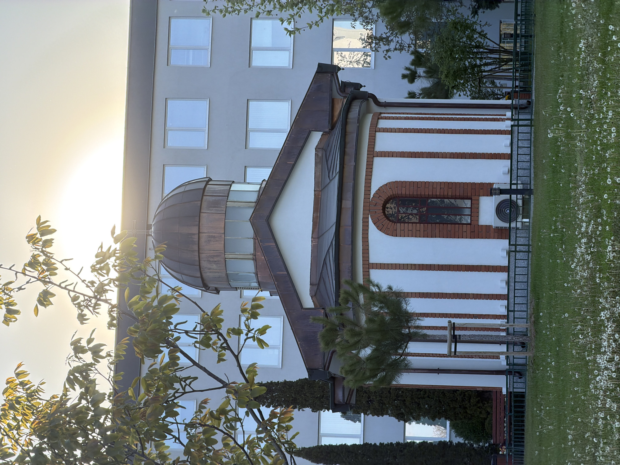 The image shows a church-like structure with a copper dome in Sofia, Bulgaria. The building has white walls with horizontal red brick stripes and an arched window. A modern building is behind it with multiple windows. The foreground features greenery, including trees and a lawn filled with dandelions. The image is taken with the sun shining brightly in the background, causing a glare. A fence separates the building from the lawn. An air conditioning unit is visible on the side of the building.