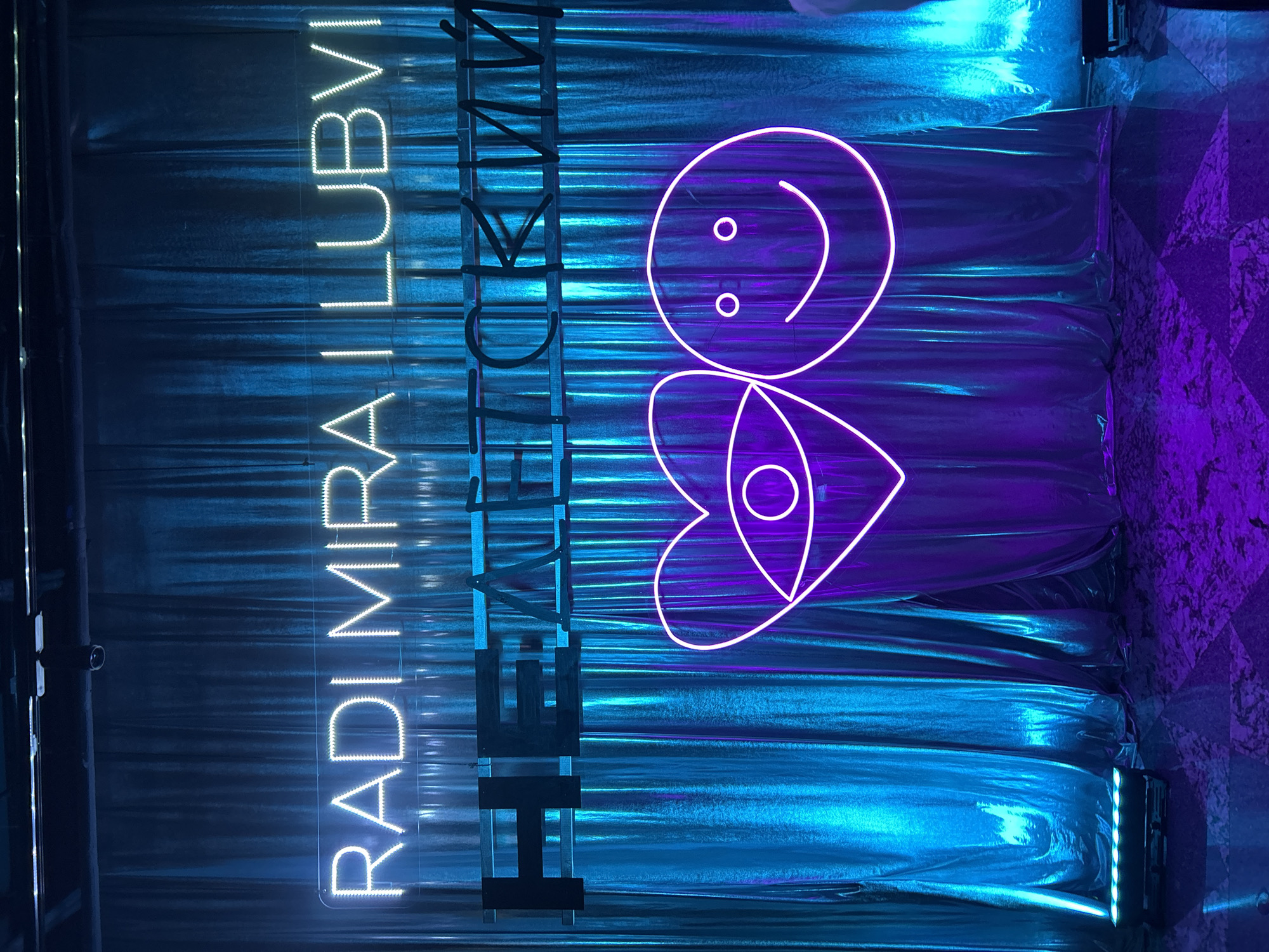 The image shows an art installation in Moscow, Russia. The installation features neon signs against a backdrop of shimmering, metallic blue and purple fabric. The signs consist of the vertically stacked words "RADI MIRA ILLUBVI" in white neon, partially obscured by black scaffolding. To the right of the text is a stylized neon drawing of a smiley face above a heart with an eye in the center, glowing in purple neon. The fabric backdrop has horizontal creases, suggesting it's draped. The lighting creates vibrant contrasts between the blue and purple areas of the fabric. The scene is likely indoors, and possibly part of a public exhibition. A portion of a patterned wall can be seen on the right edge of the image.