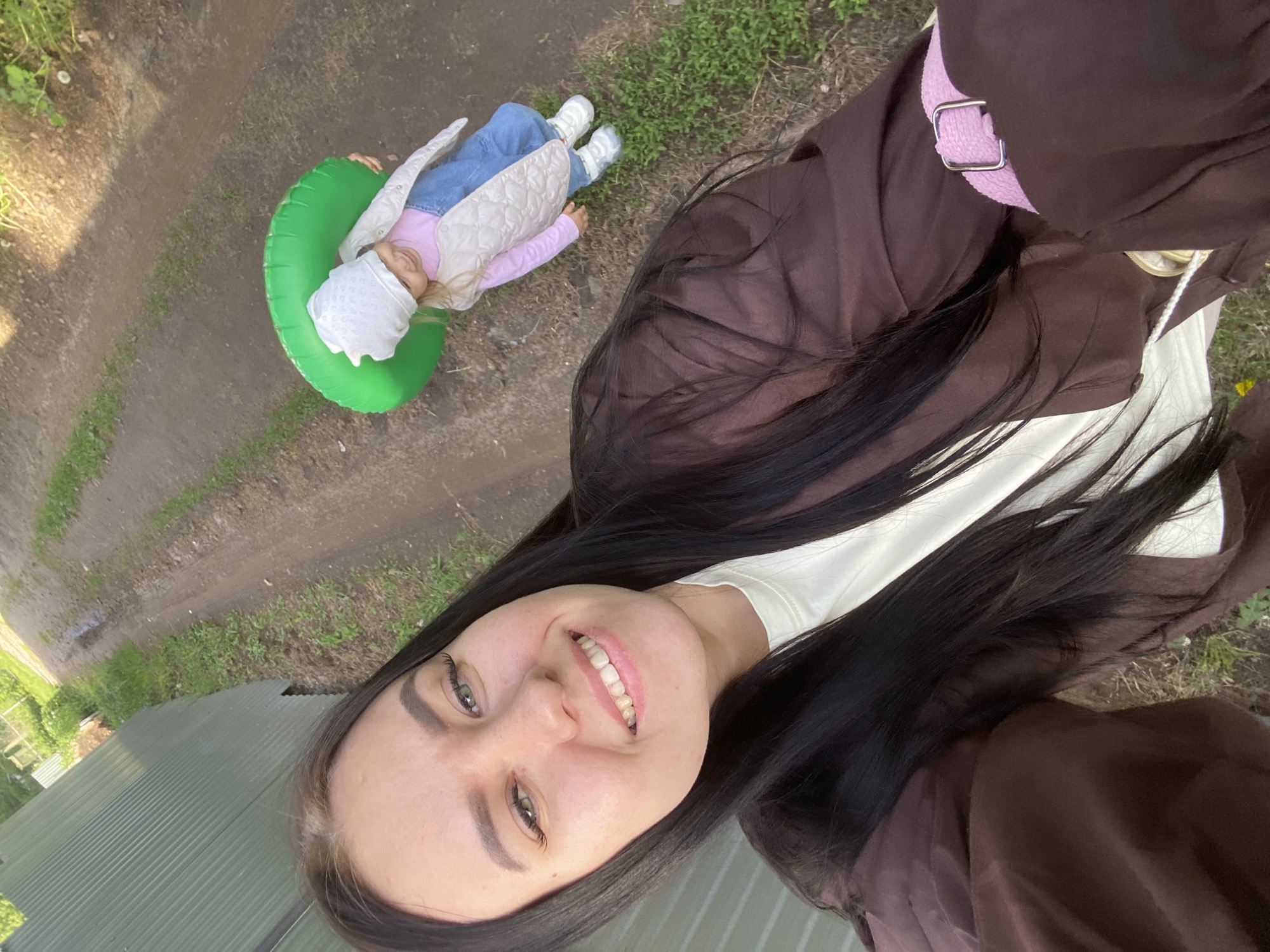 A selfie taken outdoors shows a smiling woman with dark hair wearing a brown jacket with a pink strap. In the background is a child lying on the ground in a green inflatable ring. The child is wearing a white hat, a light pink shirt, a quilted vest, blue jeans, and white shoes. The setting appears to be a grassy area with a dirt path, possibly a park. A corrugated metal structure is visible in the foreground. Given the mention of the city of Chaikovsky, Russia, it's plausible the scene is in or near a park or recreational area within that city, though no unique identifiers can confirm this.