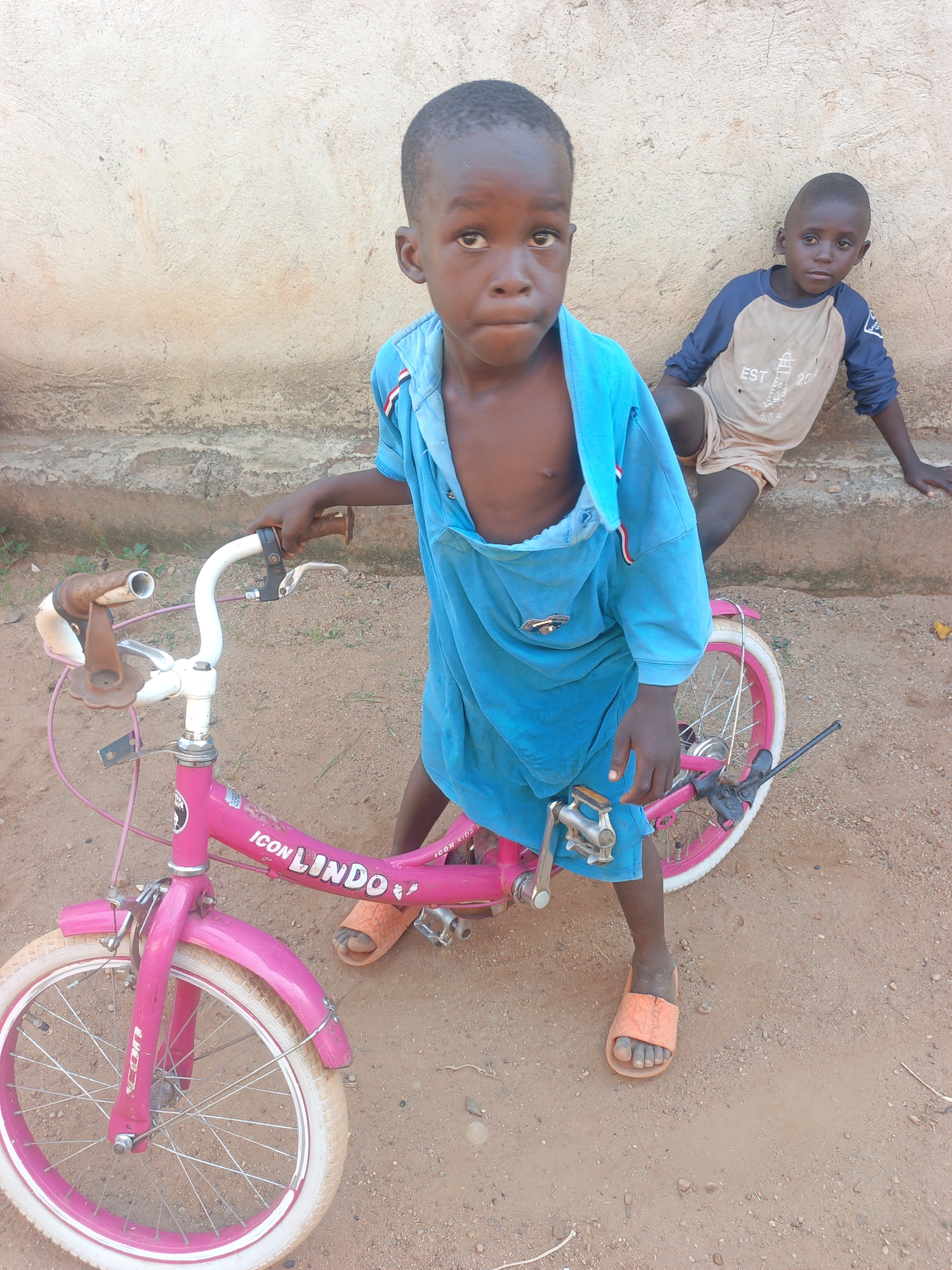 Two young boys are present in an outdoor setting in Akwanga, Nigeria. One boy, centered in the image, stands beside a pink bicycle labeled "ICON LINDO". He wears a blue, hooded shirt that is partially open, and orange sandals. The bicycle is in a used condition, with visible wear and tear on the handlebars and frame. The second boy leans against a beige wall in the background. He wears a long-sleeved shirt and light-colored shorts. The ground is dirt.