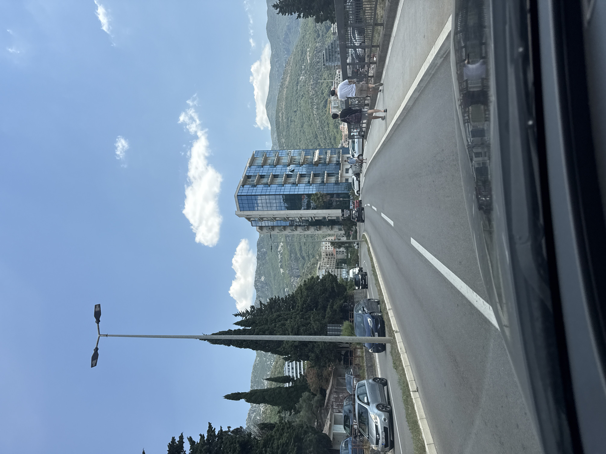 The image captures a street scene in Boreti, Montenegro, on a clear, sunny day with a few white clouds dotting the bright blue sky. The perspective is from inside a vehicle, looking forward.
The foreground shows the dashboard and windshield of the car, with the reflection of buildings and the sky visible on the windshield. The road ahead is asphalt, with white lane markings.
On the left side of the road, there are several cars parked and driving. One silver hatchback is prominently in view, followed by a dark blue hatchback. Further down the road, more vehicles are visible, including a white van and a black car. To the left of the road, there are mature cypress trees and smaller buildings, one with a red roof.
Dominating the midground is a modern, multi-story building with a glass facade, reflecting the surrounding environment. This building is situated on the right side of the road. Adjacent to this building is a sidewalk where two individuals are walking away from the camera. The person in front is wearing a black t-shirt and shorts, carrying a backpack. The person behind them is wearing a white t-shirt and shorts. A metal railing runs along the edge of the sidewalk.
In the background, lush green mountains rise, partially covered with trees. The overall environment suggests a coastal or mountainous urban setting, consistent with the location of Boreti, Montenegro. A street light with two fixtures stands tall on the left side of the road. The lighting indicates it is daytime, likely late morning or early afternoon, due to the shadows cast.