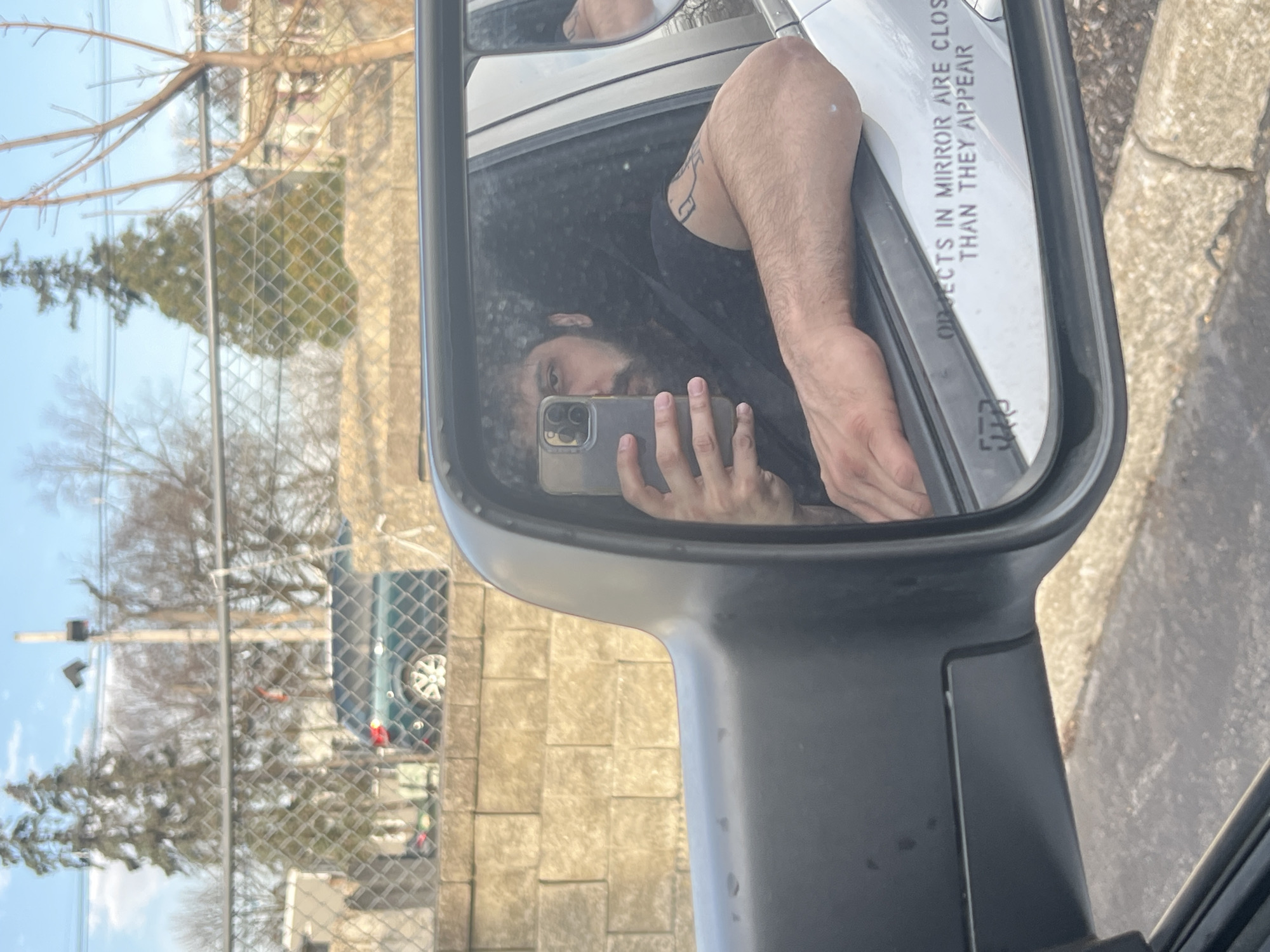 A reflection in a car's side mirror shows a person holding a smartphone, presumably taking a selfie. The person has long, dark hair and is wearing a black shirt. Part of their arm is visible, revealing a tattoo. The mirror's surface includes the warning, "OBJECTS IN MIRROR ARE CLOSER THAN THEY APPEAR." Outside the car window in the background is a chain-link fence, trees, and what appears to be a dark-colored SUV or truck. The backdrop, characterized by the fence, trees, and parked vehicle, may potentially represent an urban or suburban area within Newburgh, New York. A traffic light is also visible through the trees.