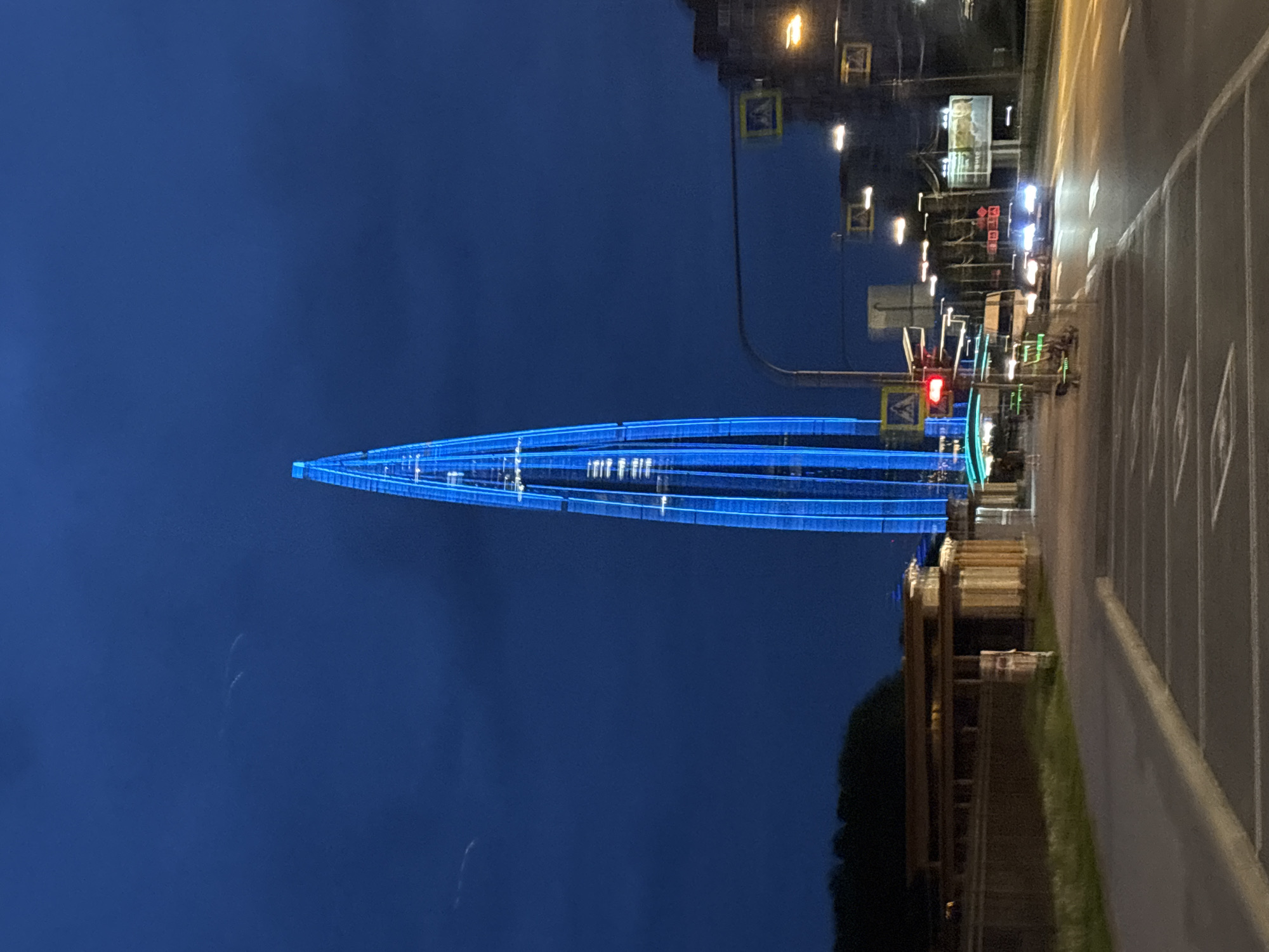 The image shows the Lakhta Center skyscraper in Saint Petersburg, Russia, illuminated with blue lights at night. The building's distinctive spire is the focal point, stretching towards the dark blue sky. To the right of the building, a road with lane markings and street lights runs along the edge of the frame. Traffic lights, one showing red, are visible along the road. In the foreground, there is some greenery and what appears to be a building or structure with visible architecture. The overall environment suggests an urban setting during the evening.