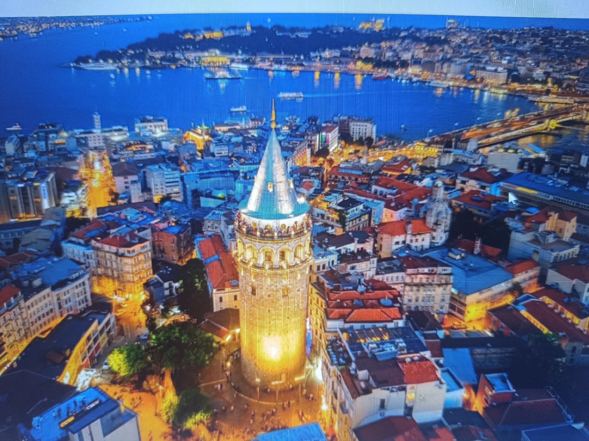 The image shows an aerial view of the Galata Tower in Istanbul, Turkey, at night. The tower is illuminated with bright golden light, standing prominently amidst a dense urban landscape. Buildings with red and blue roofs surround the tower, and streets are lit with warm yellow light. In the background, a large body of water reflects the city lights. Further beyond, the outline of a distant shoreline with more city lights is visible. There is no evidence of the city of Seewald, Germany in the image.