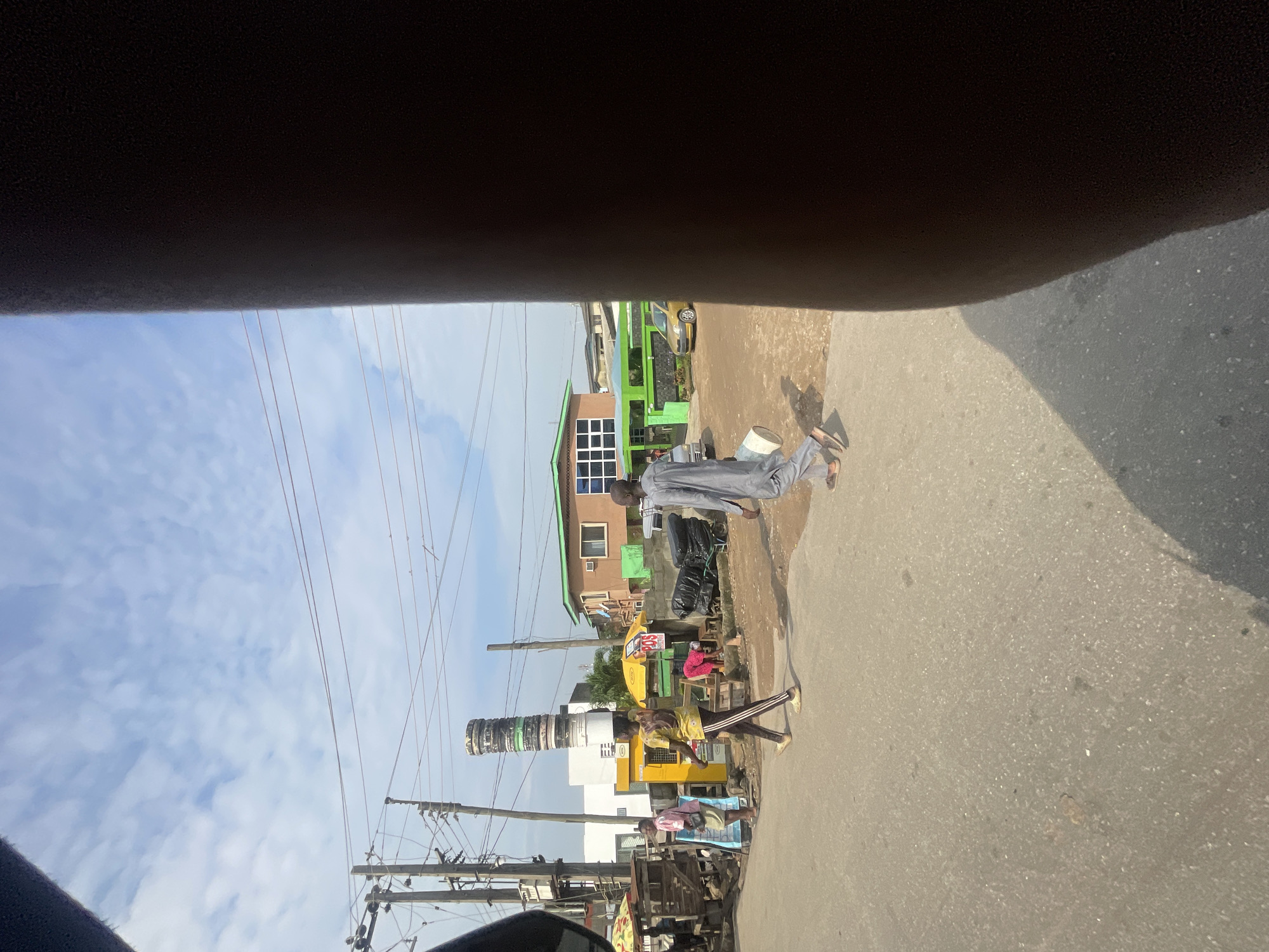 In Akowonjo, Nigeria, a street scene unfolds under a cloudy sky. The foreground is dominated by a blurred, dark object on the right, likely a person's body or clothing obscuring the view. In the middle ground, the paved road shows signs of wear. To the left of the road, a young person is walking, carrying a tall stack of buckets on their head. They are wearing a yellow t-shirt with black striped pants. Further back, a man in a grey robe is walking to the right, holding a white bucket. A few market stalls with umbrellas are visible, along with various buildings, including one painted green with white-framed windows. Utility poles with numerous wires crisscross the scene, a common feature of Nigerian urban landscapes. A yellow car is parked on the right side of the road, near a green fenced area. The overall impression is of a bustling street in a Nigerian city.