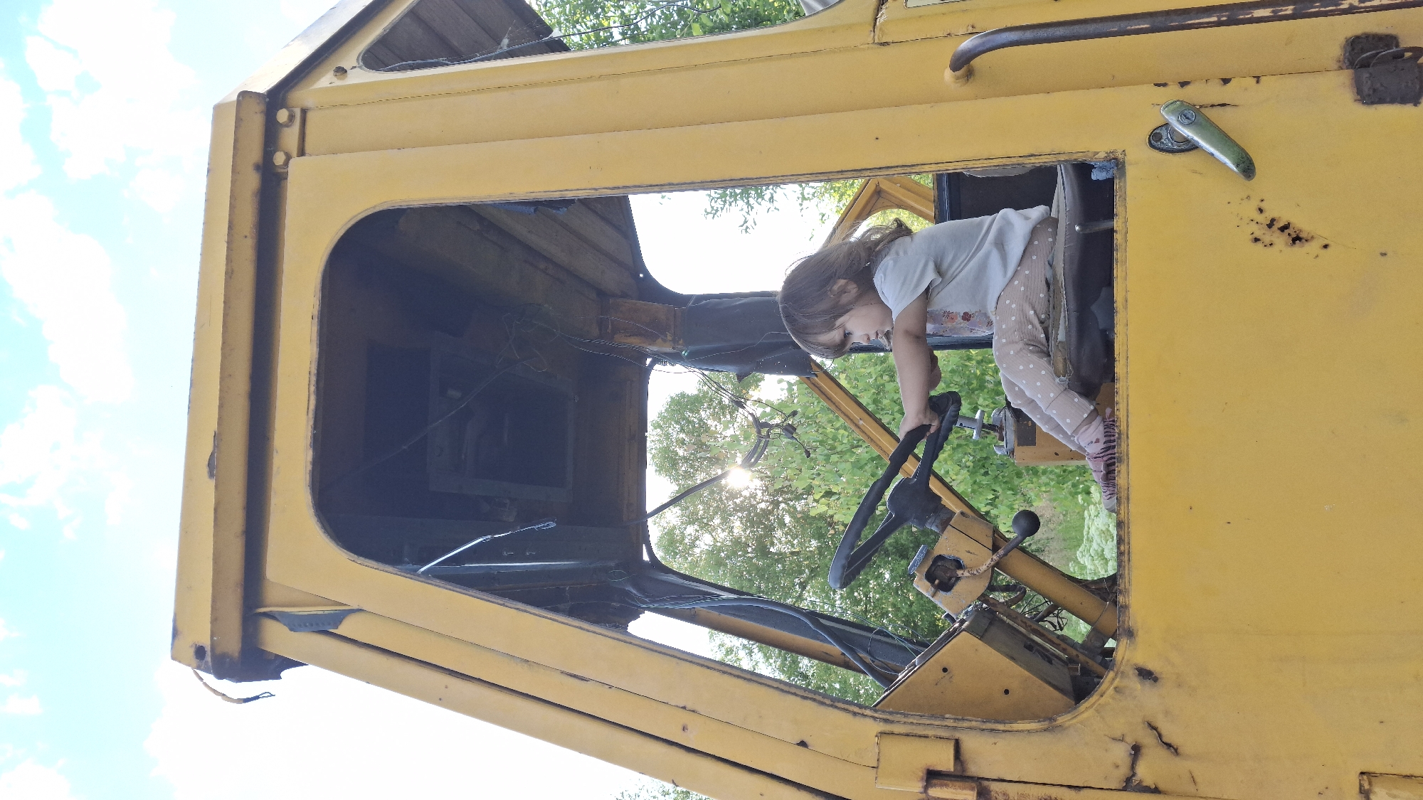 A young girl with brown hair is playing inside the cab of a weathered, yellow truck. She is leaning forward, her hands on the steering wheel, as if she is driving. She wears a light-colored t-shirt with a floral print and light-colored pants with white polka dots and pink stripes on the bottom. The truck's interior appears to be old and somewhat dismantled, with exposed wires and a broken window frame. Sunlight streams through the open windshield, creating a lens flare. Outside the truck, lush green trees and a bright blue sky with scattered clouds are visible, suggesting it is daytime and the weather is clear. The scene conveys a sense of childhood play and imagination in an outdoor setting. The location is Innolovo, Russia. There is no visible text in the image.