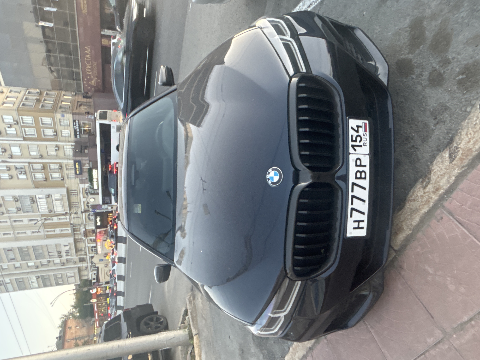 The image shows the front of a black BMW sedan parked on a city street in Novosibirsk, Russia. The car has a license plate that reads "H777BP 154 RUS". In the background, there are several multi-story buildings, a bus, and other vehicles on the road, suggesting a busy urban environment. The sky is overcast, indicating it might be daytime. The car is parked on the side of the road, with a sidewalk and a curb visible in the foreground. The scene appears to be a typical street view in a Russian city.