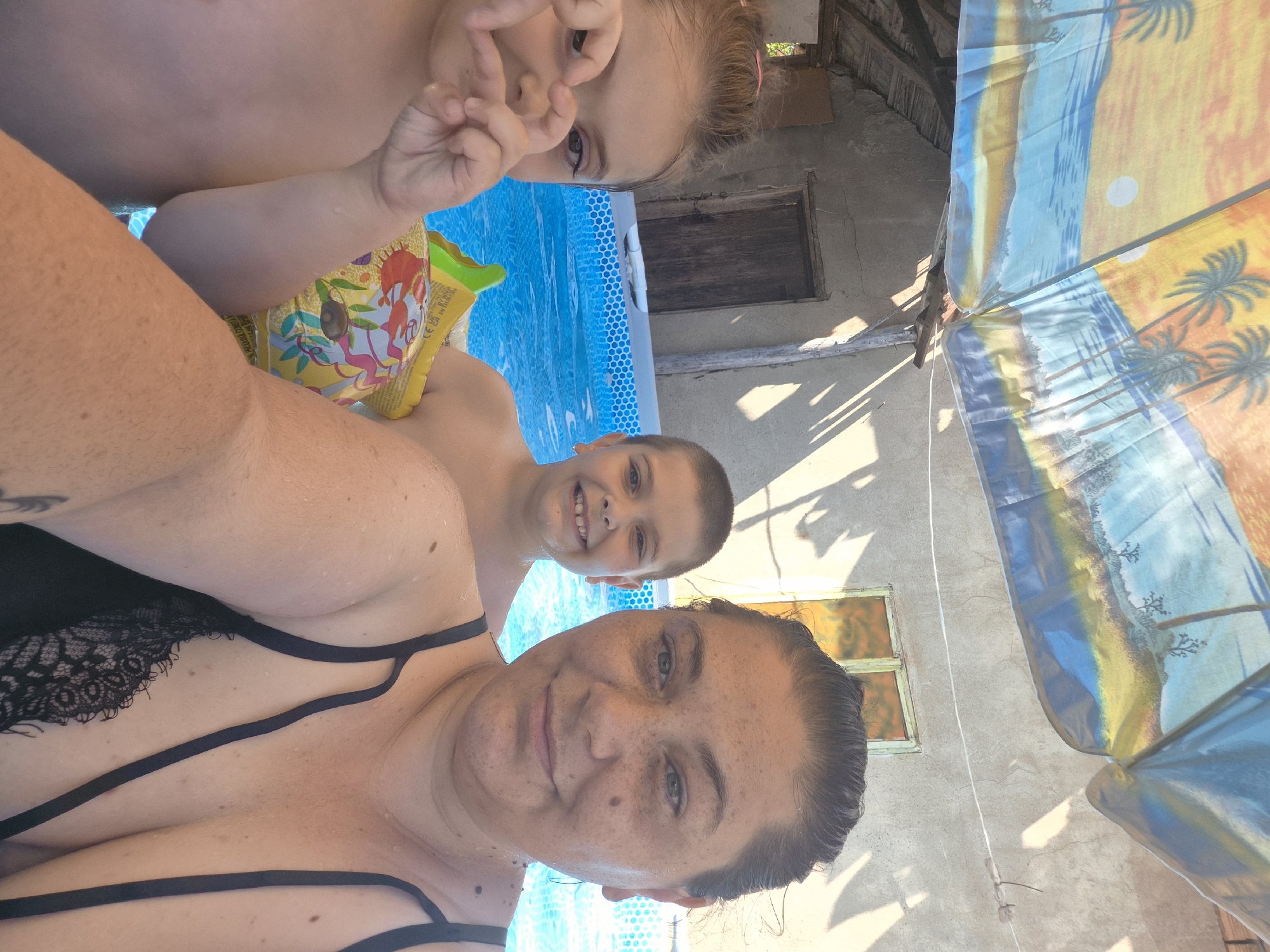 In Kalipetrovo, Bulgaria, three people are posing for a selfie in a swimming pool. On the left, a young girl makes a heart shape with her hands over her face, partially obscuring it. To her right, a young boy smiles, his head and shoulders visible above the water. The woman on the far right is in the foreground, looking at the camera with a slight smile. Behind them, a building with a rustic wooden door and a weathered facade is visible. A large umbrella with a tropical beach scene is overhead, casting shadows. The swimming pool is filled with clear blue water, and a yellow inflatable arm band with cartoon characters is visible. The overall scene depicts a casual, outdoor recreational setting on a sunny day.