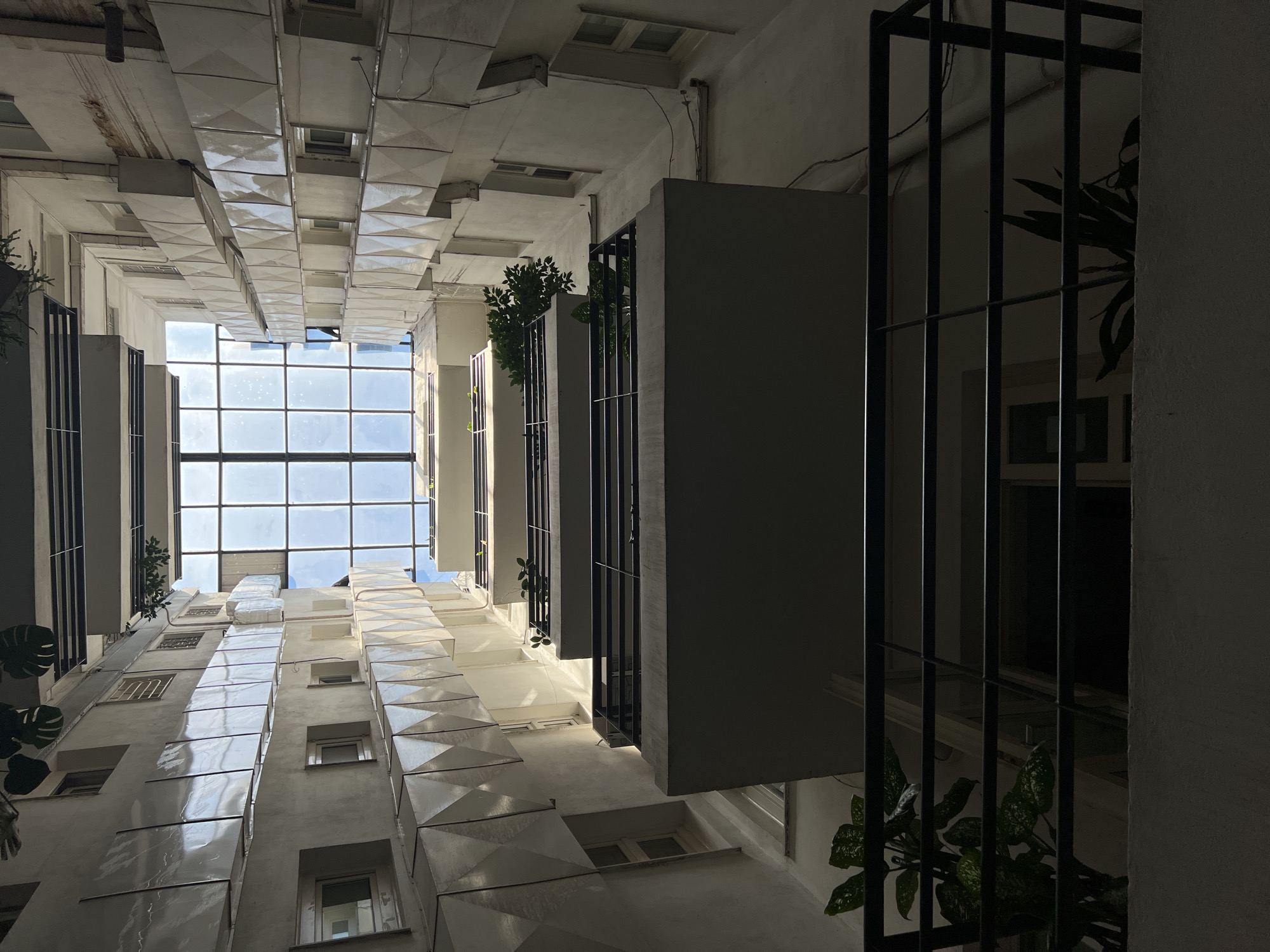 This image shows a view looking straight up from the bottom of a building's interior courtyard. The courtyard is enclosed by the walls of the building, which are lined with multiple stories of balconies and windows. The walls are a light, neutral color, possibly concrete or stucco, and feature decorative panels on the exterior facade. There are several balconies visible, each with dark metal railings. Plants are present on some of the balconies, adding touches of greenery. Above the courtyard, a large, grid-like glass roof allows sunlight to enter. The sky is visible through the glass roof, appearing bright blue with some cloud cover. The building appears to be a residential or office building, likely located in a European city. The architectural style suggests it could be in Odesa, Ukraine, given the context. There are no people visible in the image. The overall impression is one of an urban architectural space.