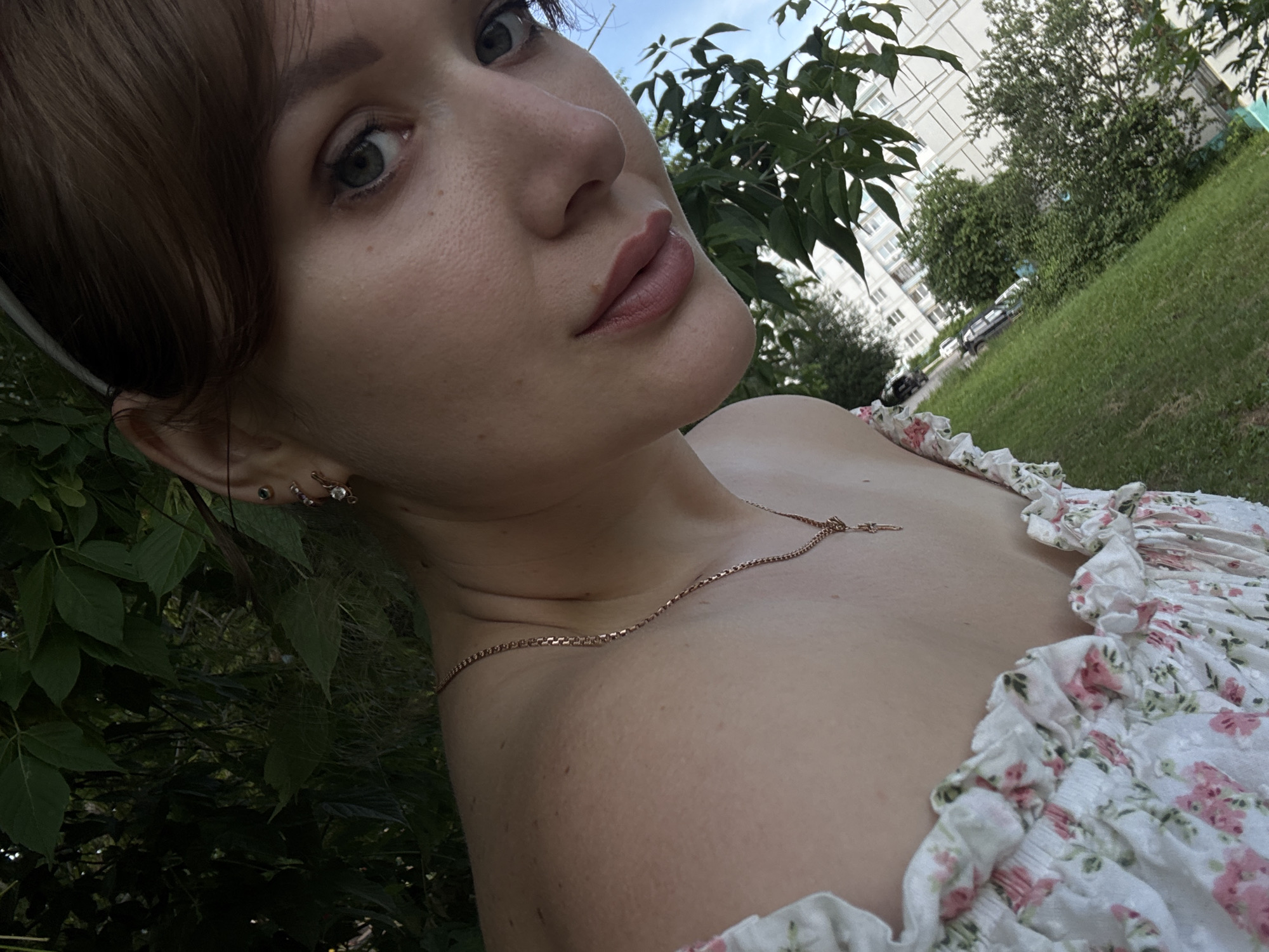 The image shows a close-up, low-angle selfie of a young woman outdoors, likely in a residential area of Chaikovsky, Russia, given the context provided by the user. The woman has light skin and brown hair, and is wearing a white dress with a floral pattern. She is adorned with subtle makeup and jewelry, including a delicate necklace and earrings. Her face is partially obscured by foliage, suggesting she is near trees or bushes. In the background, a grassy area is visible, with parked cars and apartment buildings, characteristic of urban landscaping in Russia. The sky is blue and clear, indicating fair weather. The overall scene depicts a casual moment outdoors, possibly in a park or courtyard.