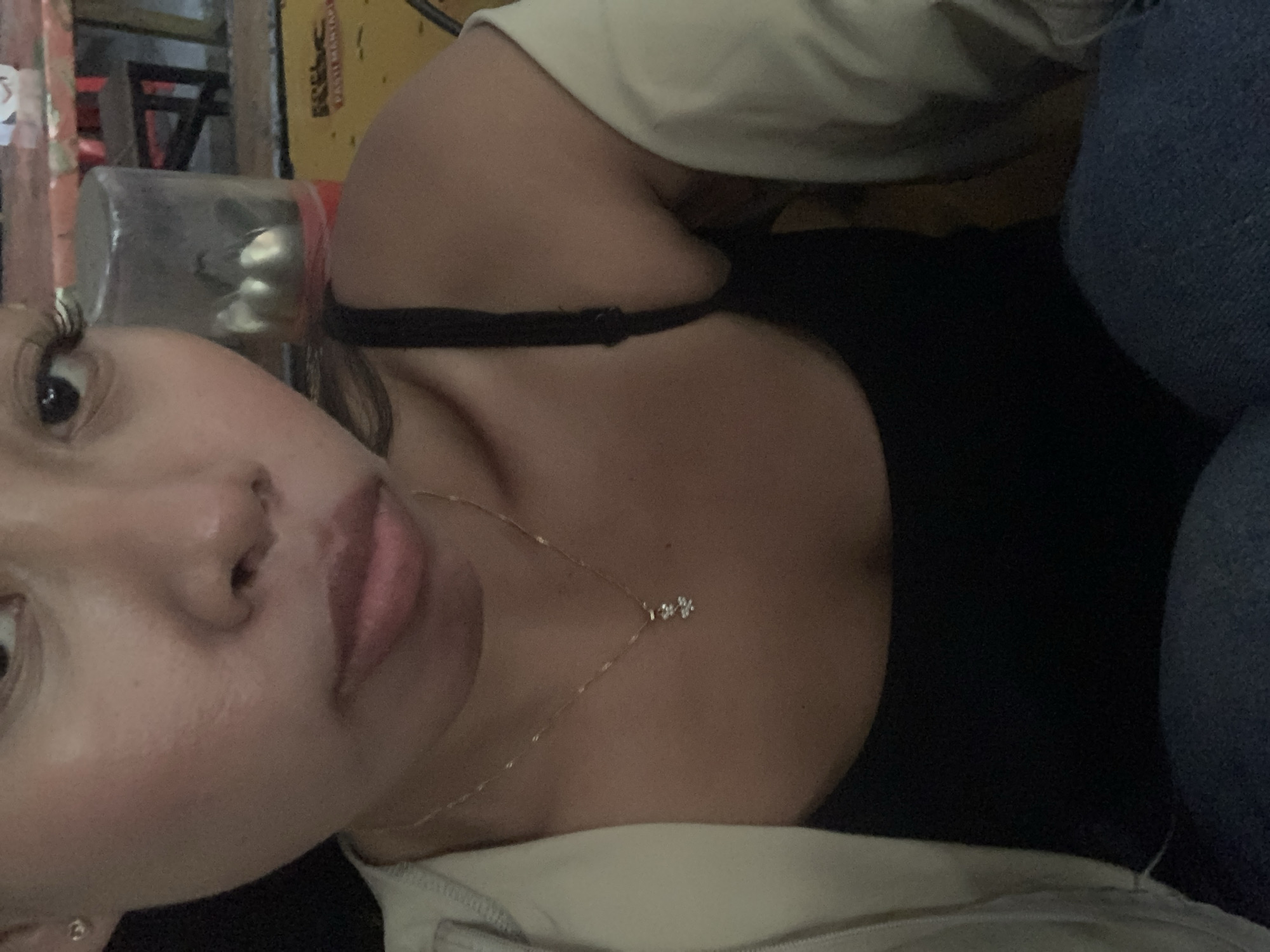 A close-up, high-angle selfie shows a young woman with fair skin, dark hair, and subtle makeup, including mascara and lip gloss. She is wearing a black tank top and a light-colored jacket draped over her shoulders. A delicate gold necklace with a small charm is visible on her neck. The background is blurred, but appears to be an indoor setting with yellow and red accents, possibly a restaurant or cafe. The overall lighting is dim, creating a warm and intimate atmosphere. The image is likely taken in Tamanbaru, Indonesia, given the user's prompt.