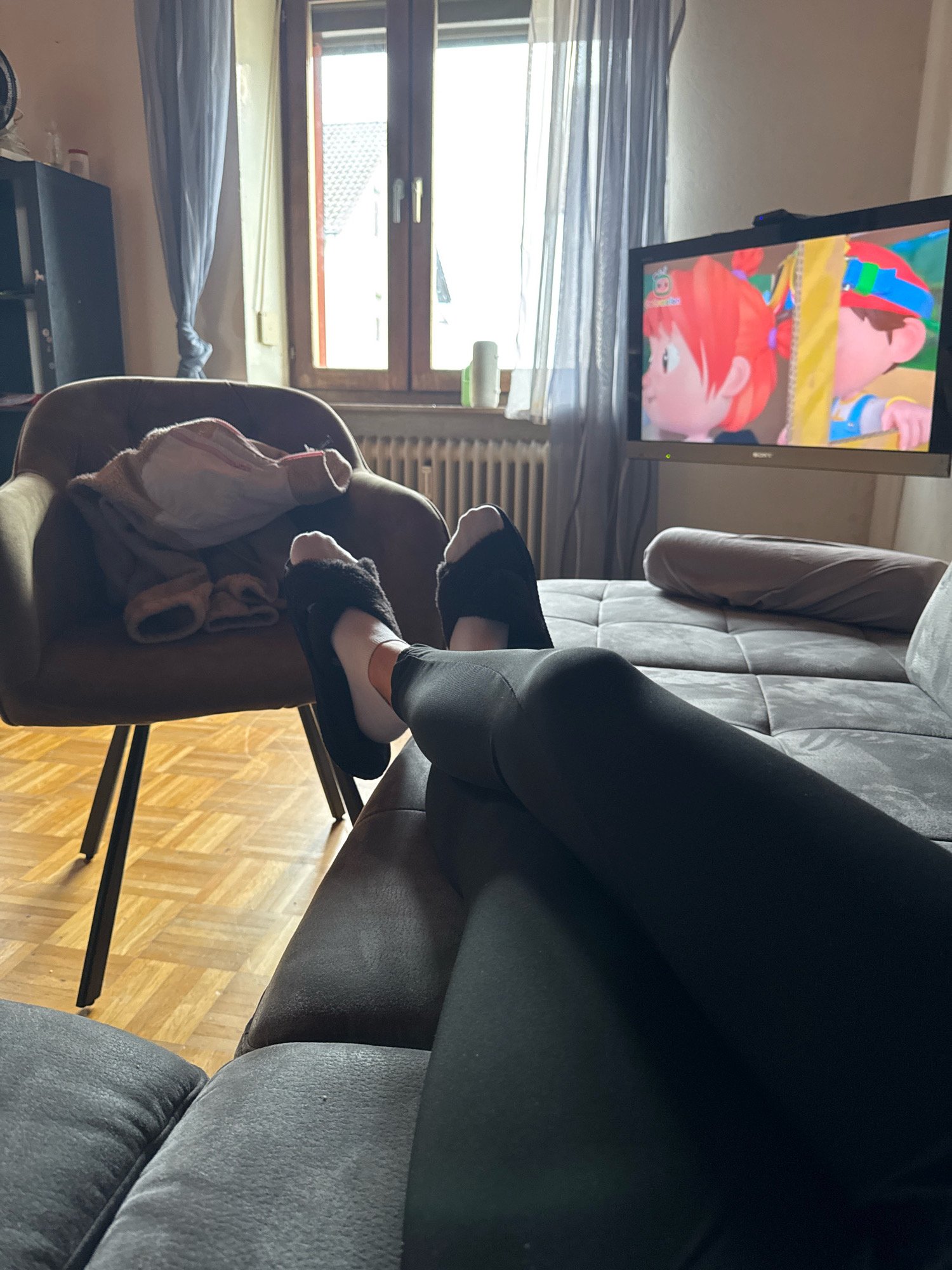 The image depicts an indoor scene within a residential apartment, likely in Überherrn, Germany. The focal point is a person's legs and feet, clad in black leggings and black fuzzy slippers, resting on a dark gray sectional sofa. The person's feet are positioned towards the right side of the frame.
To the left, a brown armchair is visible with some clothing draped over it, appearing to be a jacket and a pair of pants or a blanket. The armchair has dark metal legs.
In the background, a television set is mounted on a stand, displaying a colorful cartoon featuring animated children. The TV is positioned to the right of the armchair.
Behind the armchair and to the left of the TV, a window with light blue curtains is present. The window shows a view of what appears to be houses with pitched roofs and a cloudy sky. A radiator is visible beneath the window.
The flooring in the visible area is parquet flooring laid in a herringbone pattern. The overall environment suggests a casual, relaxed setting, possibly a living room. The lighting suggests it is daytime, with natural light filtering in from the window.