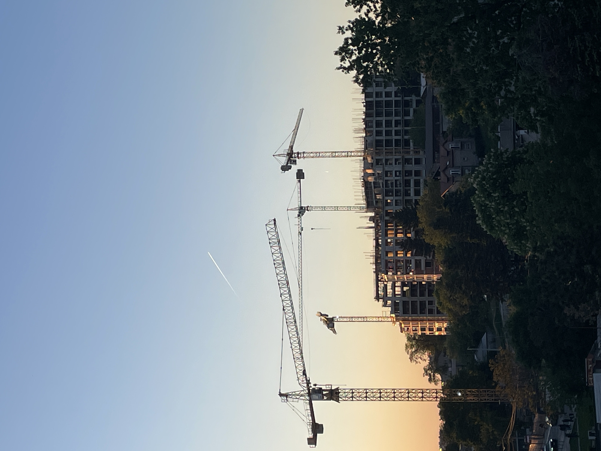 The image shows a cityscape at dusk, likely in Belgrade, Serbia, given the architectural style. Numerous construction cranes dominate the foreground, silhouetted against a sky transitioning from blue to orange. Several high-rise buildings are under construction, their skeletal frameworks visible behind the cranes. Trees partially obscure the view of other buildings and structures along the horizon line. A contrail from an airplane bisects the upper left portion of the sky. No people are visible.