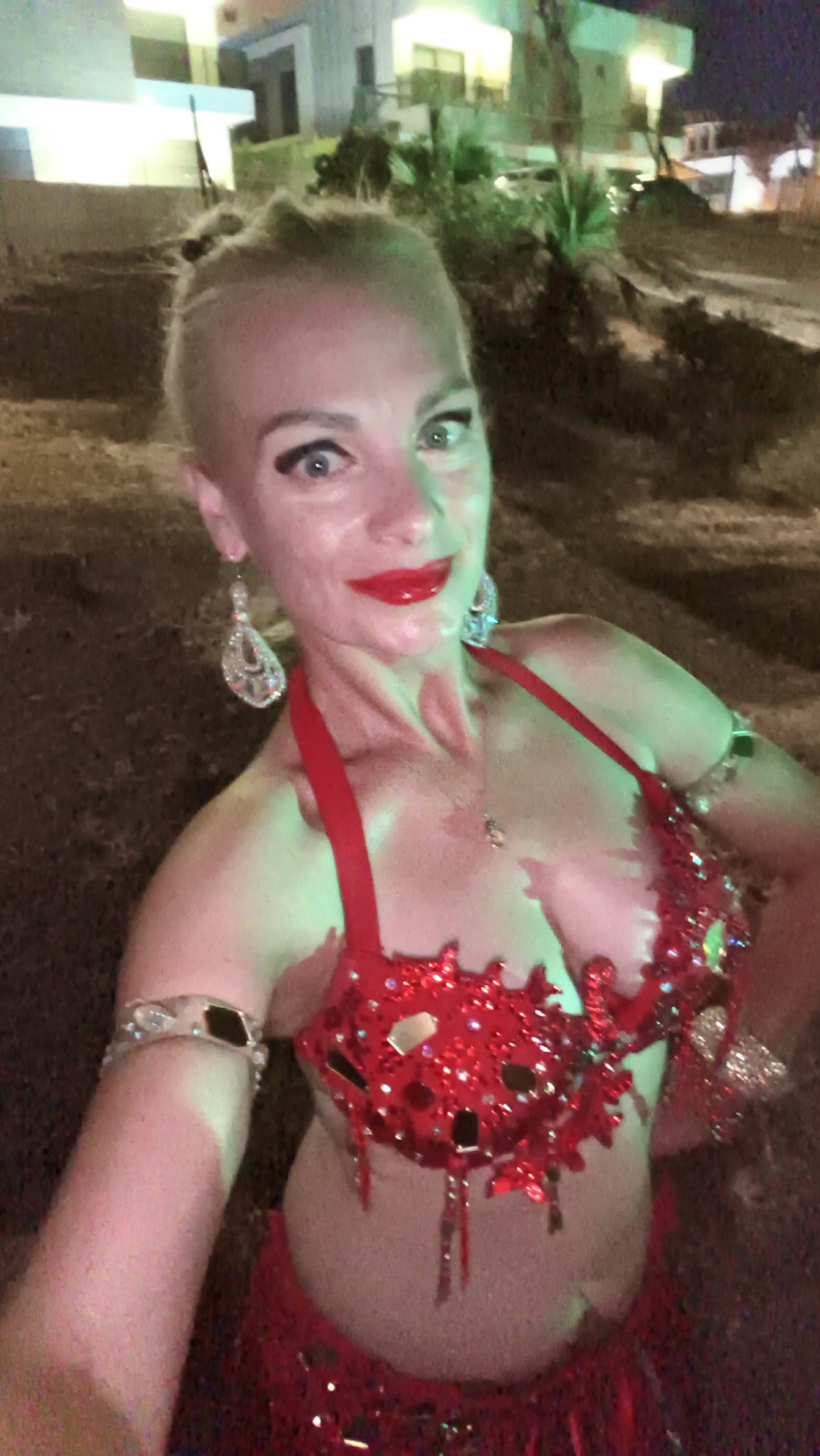 An adult Caucasian female with blonde hair pulled up in a bun, wearing a red sequined belly dancing costume, is posing for a selfie at night. The costume includes a bra adorned with sequins and dangling beads, a matching skirt, and armbands. She has bold red lipstick, visible earrings, and a necklace. The background shows a building with lights on, indicative of a residential or commercial area, with palm trees and vegetation. The scene is lit by artificial lights, casting green and white tones on the subject and the environment. The image appears to have been taken outdoors.