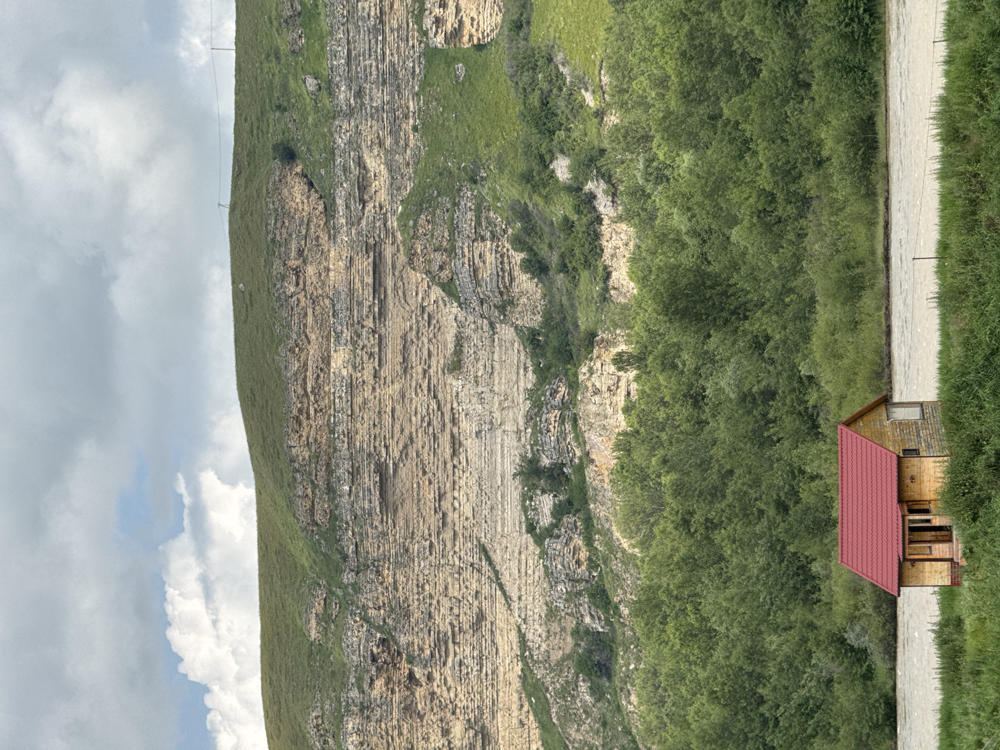 The image depicts a serene natural landscape with a small, wooden house featuring a red roof situated in the foreground. The house is located on the bank of a wide, murky river, bordered by lush green grass and dense trees. In the background, a massive, layered rock formation dominates the scene, its stratified layers indicative of geological history. The rock face is partially covered in green vegetation, and the sky above is overcast with dramatic clouds. The setting appears to be a rural or wilderness area, possibly in the vicinity of the village of Kubina, Russia, given the natural terrain and architecture. There are no people or animals visible in the image, and the environment suggests a tranquil, untouched environment.