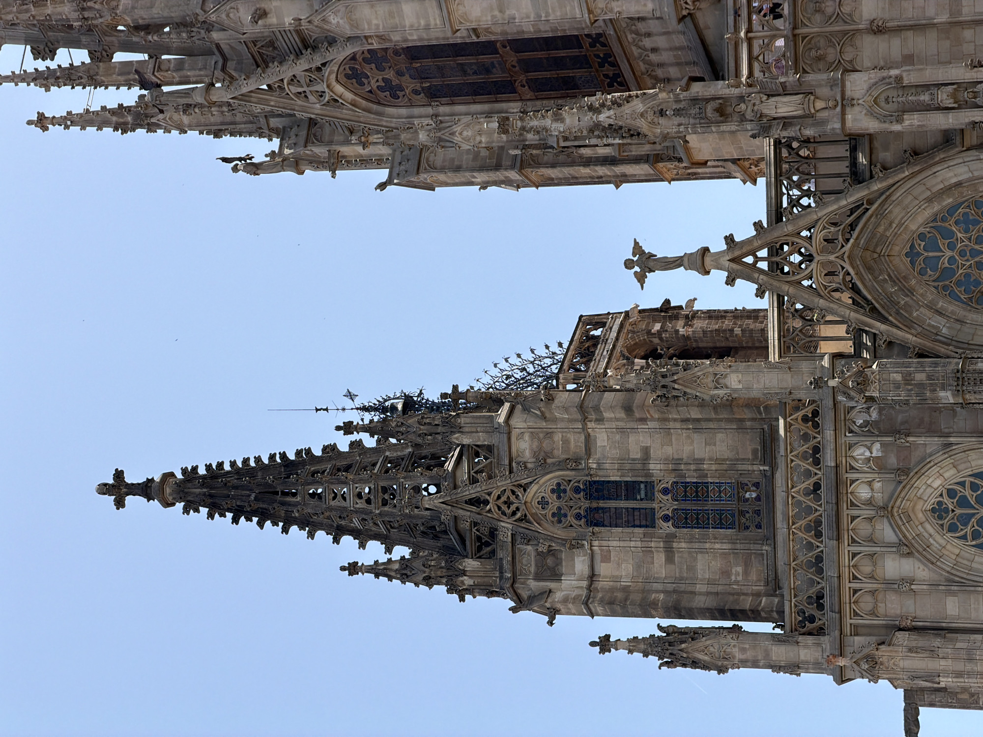 The image shows a close-up, low-angle view of the Barcelona Cathedral, a Gothic Roman Catholic cathedral located in Barcelona, Spain. The architecture is characterized by its intricate stonework, spires, and stained-glass windows. In the foreground, a statue of an angel stands atop a pointed archway. The sky is clear and blue, with a few distant birds visible. The cathedral's facade is made of weathered stone, with detailed carvings and decorative elements. The overall impression is one of grandeur and historical significance.