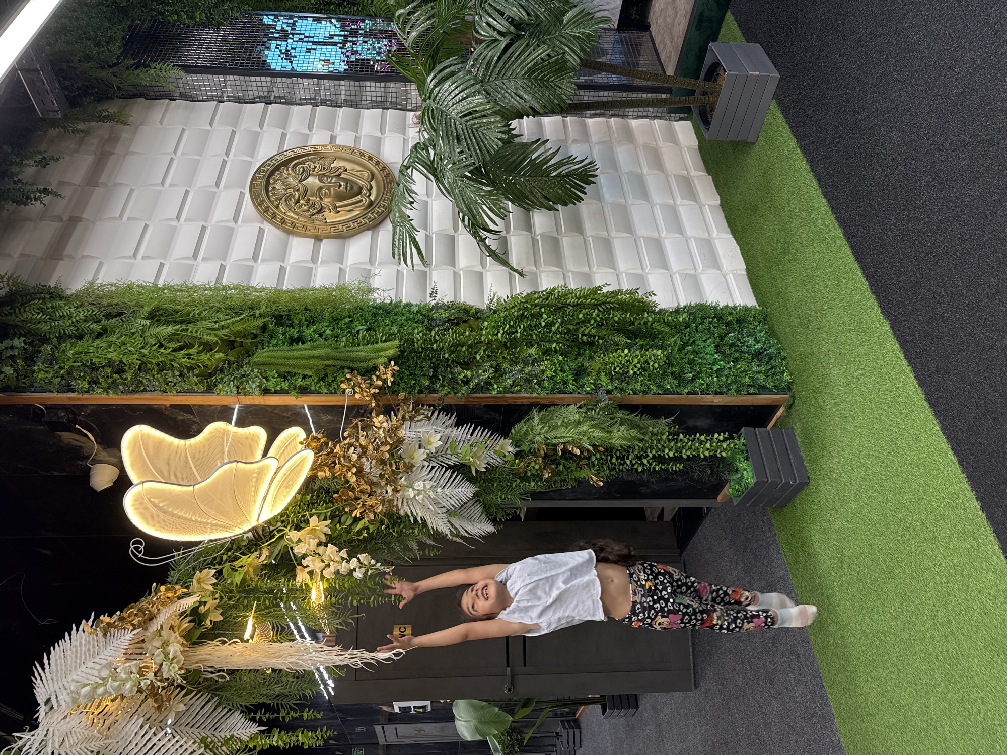 A young girl with long dark hair, wearing a white t-shirt and patterned leggings, is captured mid-jump with her arms outstretched towards hanging greenery and a large illuminated butterfly decoration. The scene appears to be indoors, possibly a decorative entryway or event space, with elements of artificial greenery and stylized wall panels. A gold medallion with a human face is mounted on a textured white wall to the right. The floor is a mix of dark carpet and artificial turf. The overall impression is one of playful interaction within a themed environment. The presence of the name "Sochi, Russia" suggests the location.