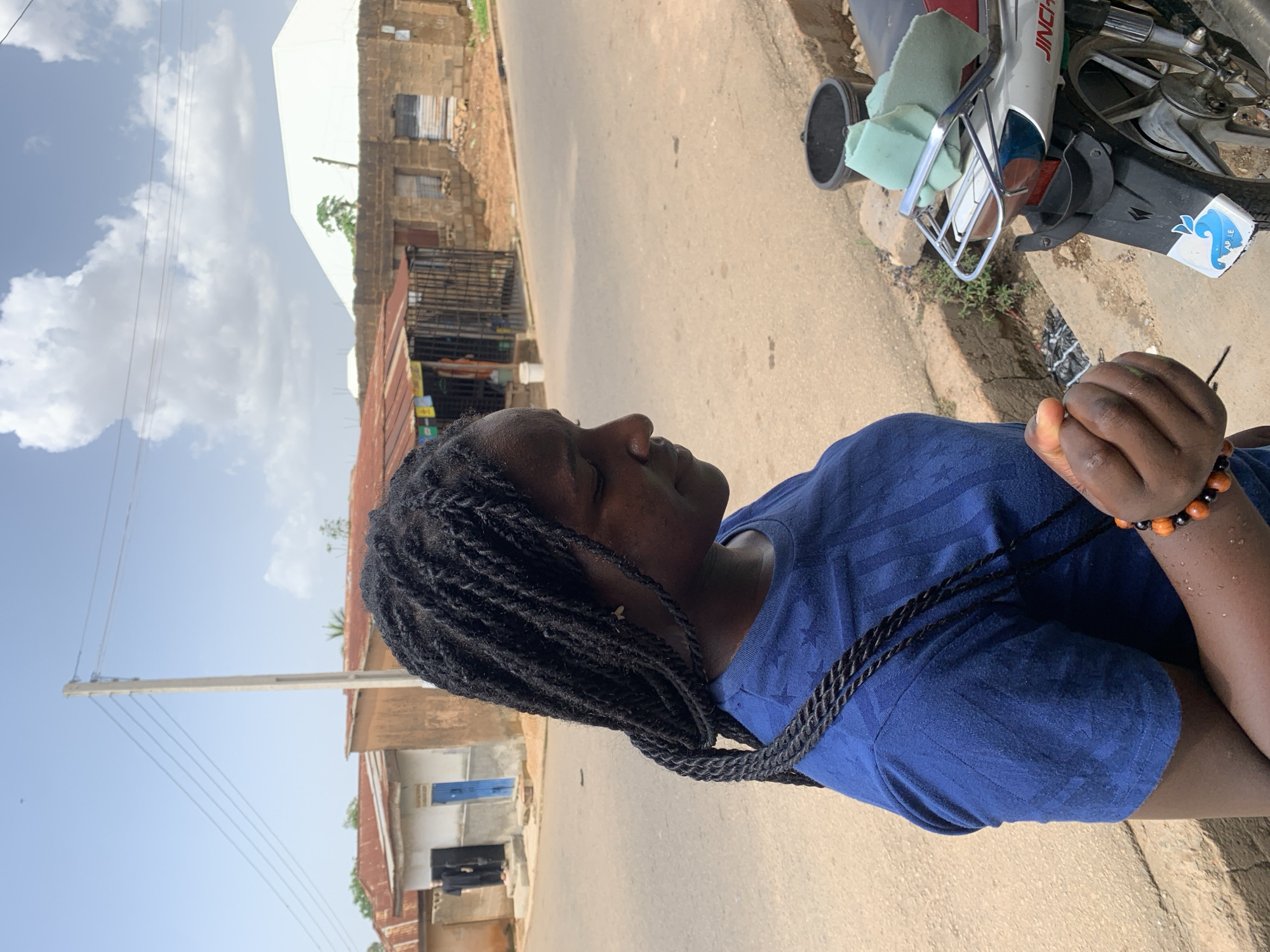 A dark-skinned woman with long, braided hair, wearing a blue t-shirt, stands on a dirt road in a city, possibly Jos, Nigeria. She is looking down, her right hand holding some stringy material. Next to her is a motorcycle, with a teal sponge on the back. Buildings with corrugated iron roofs line the street behind her. The sky is blue with scattered clouds.