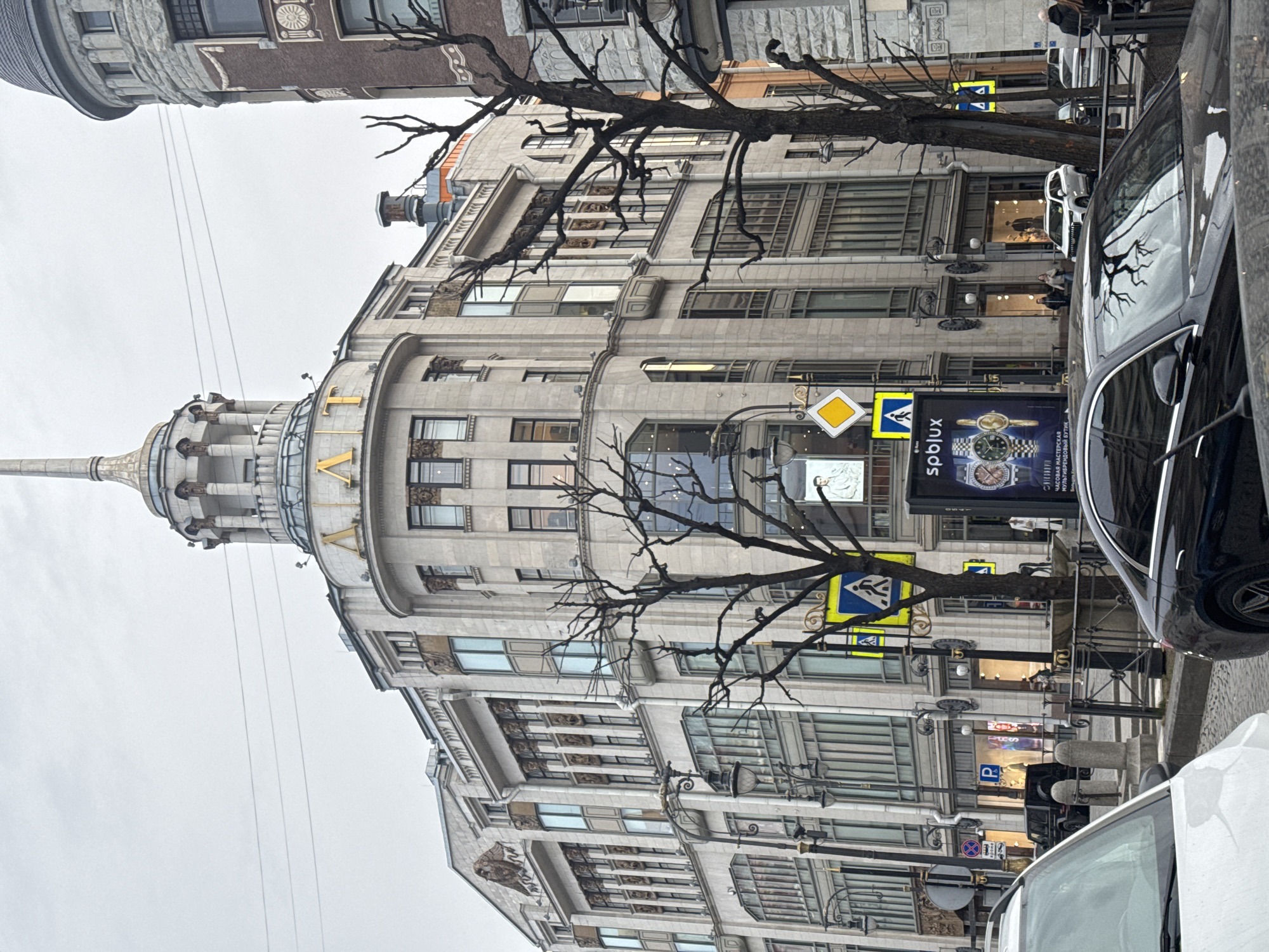 The image captures a street scene in Saint Petersburg, Russia, dominated by a large, multi-story building with a distinct architectural style. The building features intricate facade detailing, rows of windows, and a prominent tower or spire-like structure at its corner. Gold lettering is visible on the building's facade, partially obscured by the camera angle.
A bare tree, likely deciduous, stands in front of the building. Street signs are visible on the sidewalks, with pedestrian crossing symbols prominent. Several cars are parked along the side of the street. The sky is overcast. Pedestrians are visible, walking. A billboard displays a watch advertisement.