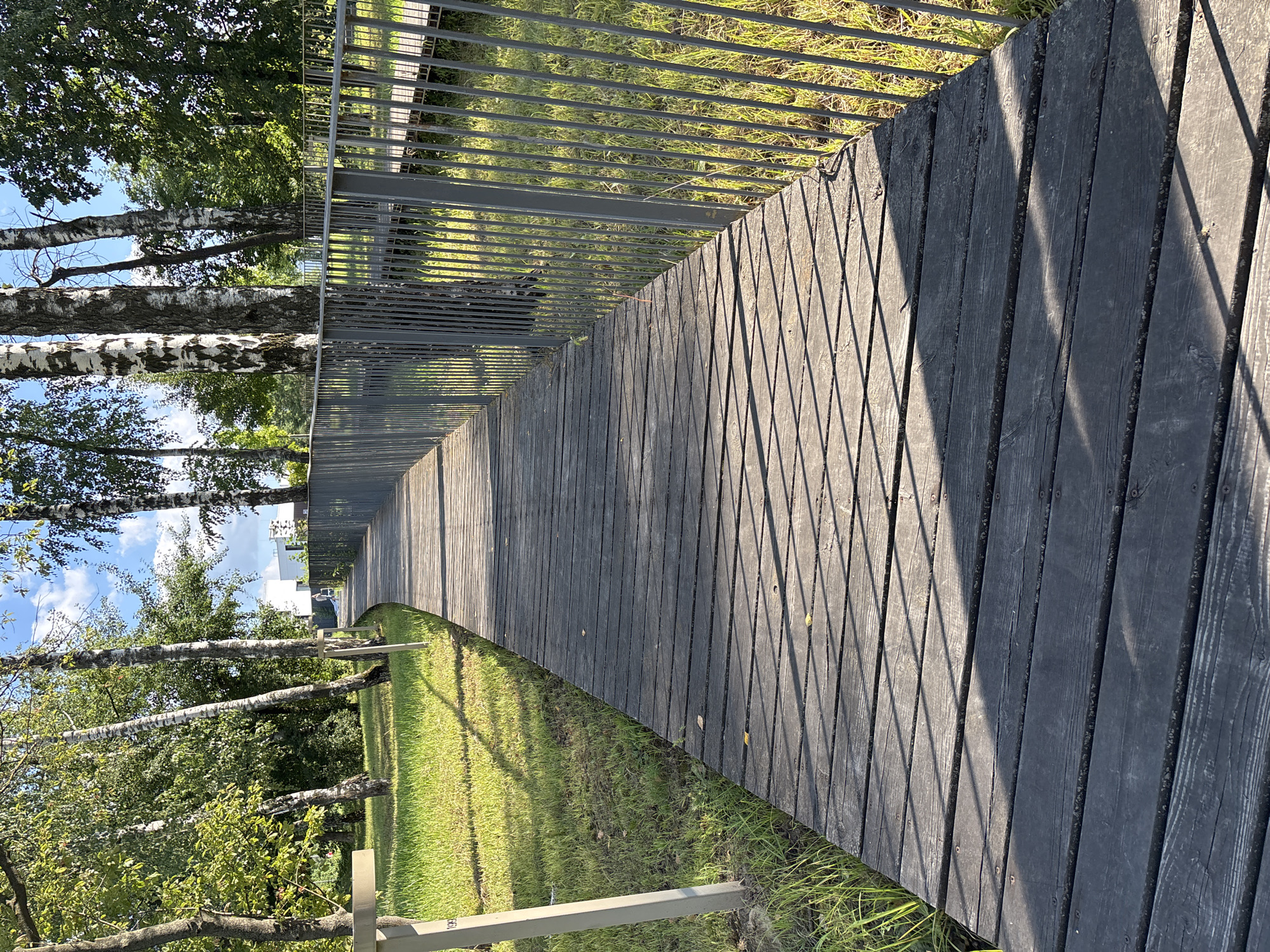 A wooden boardwalk curves through a park with lush green grass and trees. The boardwalk is made of dark, weathered wooden planks and is bordered by a metal railing. Sunlight casts sharp shadows across the boardwalk. In the distance, a modern white building is visible among the trees. The sky is blue with scattered white clouds.