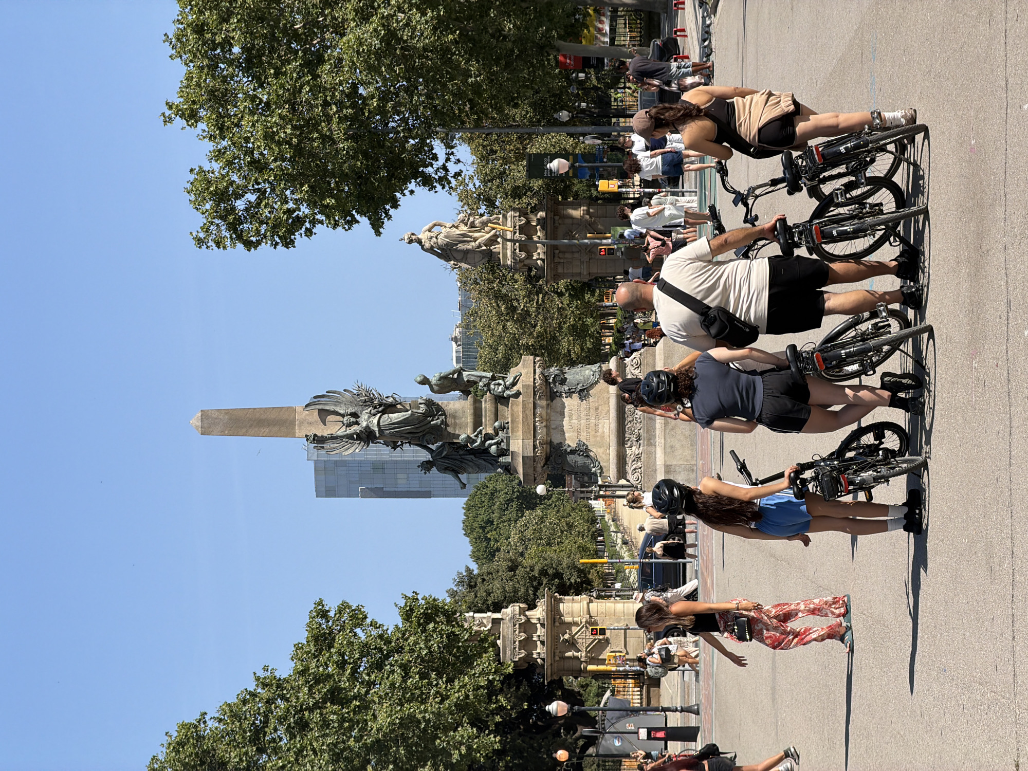 In Barcelona, Spain, a group of four people, two men and two women, stand with bicycles in an open paved area. The men are wearing t-shirts and shorts, while the women are in shorts and t-shirts. Two of the individuals are wearing helmets. The bicycles are parked upright, with handlebars interlocked. In the background, a large monument with statues and an obelisk stands prominently. Trees line the perimeter of the area, and a tall modern building is visible in the distance behind the monument. The sky is clear and blue. The scene appears to be a tourist or recreational area.