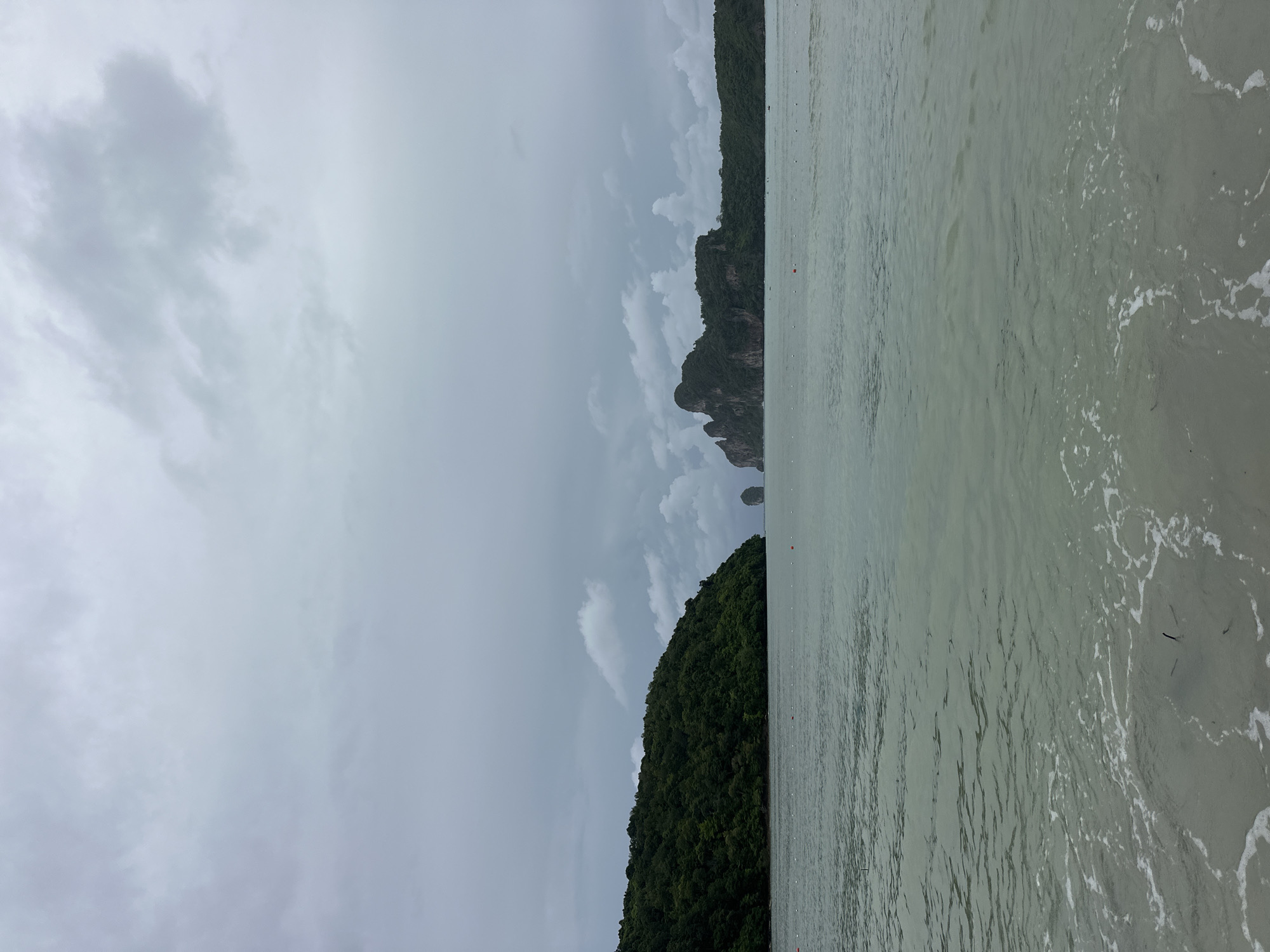 The image depicts a serene, overcast seascape with a distinctive island formation in the distance. The foreground features calm, shallow water with gentle waves lapping at a sandy shore, creating a foamy white edge. The water itself is a muted, greenish-blue hue, reflecting the overcast sky.
Dominating the middle ground are two prominent islands or rock formations. The island to the left is covered in lush, dark green foliage, with a sloping coastline meeting the water. To the right, a larger, more dramatic island rises from the sea. This island features jagged, rocky peaks that are also covered in dense vegetation, giving it a rugged appearance. Two smaller, more slender rock formations are visible in the distance between the two larger landmasses.
The sky is a vast expanse of cloudy, grey tones, suggesting a subdued light. A few scattered, wispy clouds are visible against the heavier cloud cover. The overall atmosphere of the image is one of tranquility and natural beauty. The absence of people or man-made structures, other than a few small, orange buoys visible in the water, emphasizes the pristine natural setting. This scene is consistent with the geography of Ao Nang, Thailand, known for its limestone karsts and tropical island landscapes.