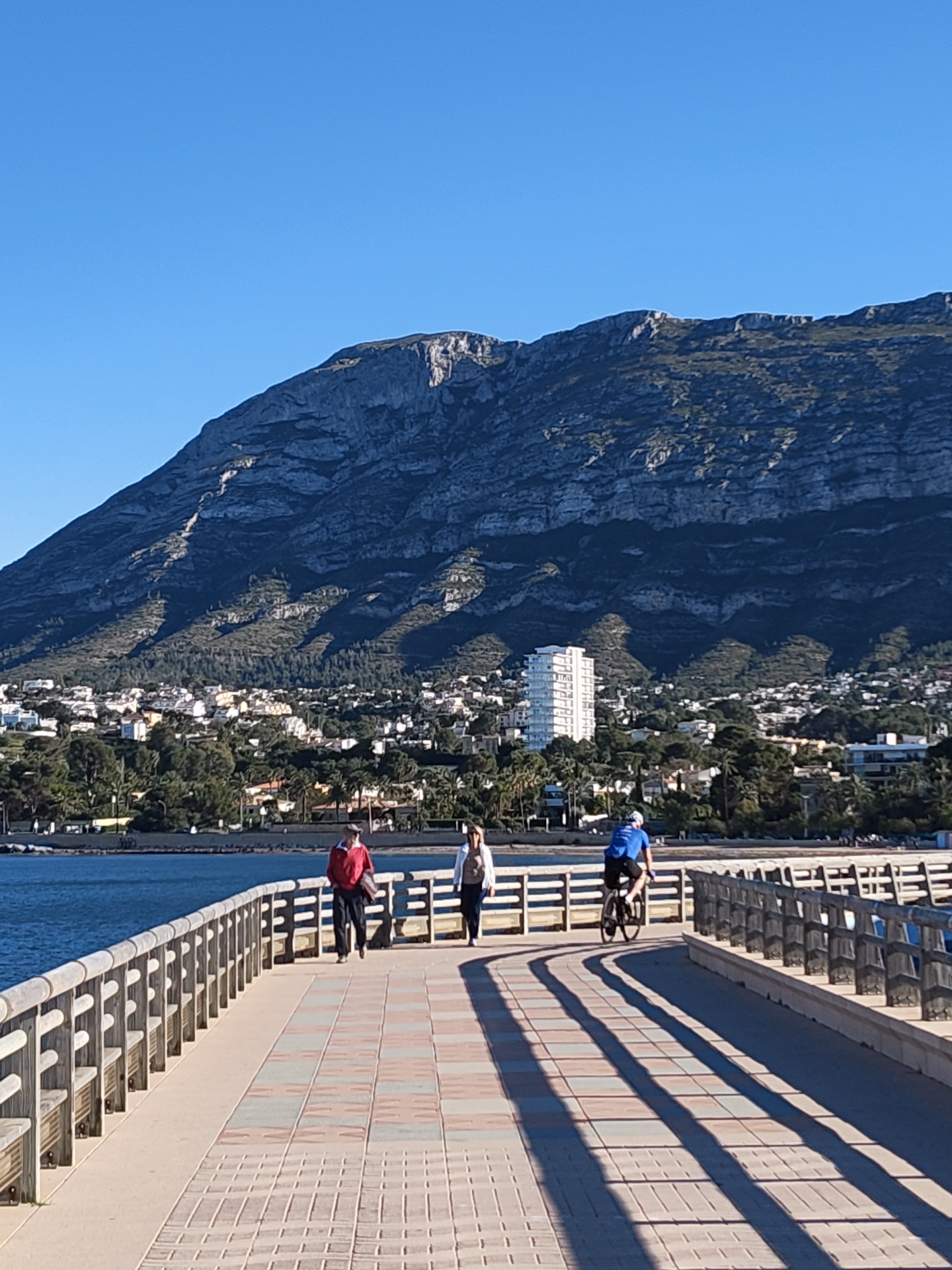 This outdoor photograph captures a clear, sunny day in Dénia, Spain, featuring a picturesque coastal landscape. The image is taken from a wide, paved pier or promenade, looking across a calm body of water towards a town nestled at the base of a grand mountain range.
In the background, a large, rugged mountain dominates the skyline, displaying distinct grey rock formations and scattered vegetation. Below the mountain, a residential area spreads out, characterized by numerous white and light-toned buildings interspersed with dense green trees, including palm varieties. A tall, modern white building stands out prominently in the middle of the town.
The foreground is occupied by the pier, paved with light-colored, patterned bricks and flanked by sturdy wooden railings on both sides. To the left, the deep blue water of the sea or bay is calm, reflecting the bright, cloudless sky above.
Three people are visible on the pier. On the left, two individuals walk towards the camera: one in a red jacket and cap, and the other in a light-colored jacket. Further along the pier, a person cycles away from the camera, wearing a blue top and a helmet. Long, dark shadows from the railings and individuals stretch across the pier, indicating a low sun angle, suggesting either early morning or late afternoon. The scene conveys a peaceful, active atmosphere in a popular recreational setting. No discernible text is visible in the image.