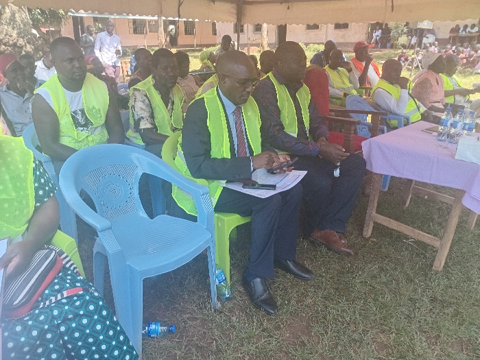 The image displays an outdoor gathering situated on a grassy area in Idakho South ward, Kenya, under a large white canvas tent. Approximately 20 to 30 individuals are visible, primarily adults, with a significant number wearing bright yellow or lime green safety vests over various casual and formal attire.
In the foreground, from the left, a person partially visible in a green vest and a dark skirt with white polka dots is seated. Adjacent is an empty light blue plastic chair. To its right, a man in a white sleeveless shirt and a yellow vest sits on a light green plastic chair, looking to the right. Next to him, a man in a dark suit with a white shirt and patterned tie, wearing a yellow vest, is seated on a light green plastic chair, looking down at a document while holding a smartphone. To his right, another man in a dark shirt and trousers, also wearing a yellow vest, is seated, holding a smartphone.
To the immediate right of the foreground individuals, a wooden table draped with a light purple cloth holds six plastic water bottles. Further back, numerous other attendees, many also in yellow vests, are seated in rows on various plastic and wooden chairs. One male attendee in the mid-ground wears a red hat. In the distant background, beyond the tent, light-colored buildings with reddish-brown roofs are visible, along with some foliage. Plastic water bottles are also observed on the ground near the chairs.